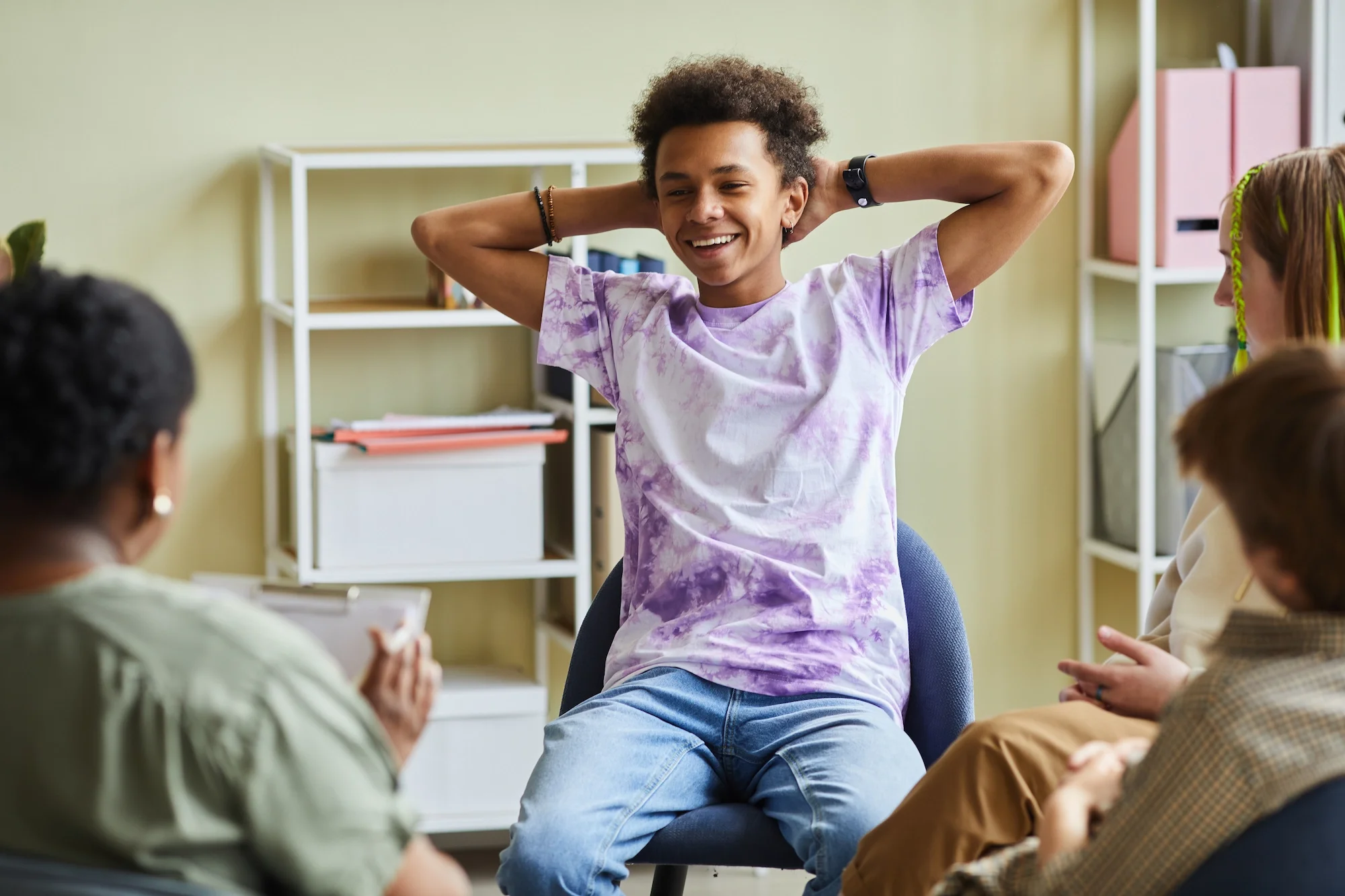 A teenager girl with curly hair, smiling, wearing a tie-dye purple shirt, sitting on a chair with her legs crossed, leading a discussion in a classroom with other students.