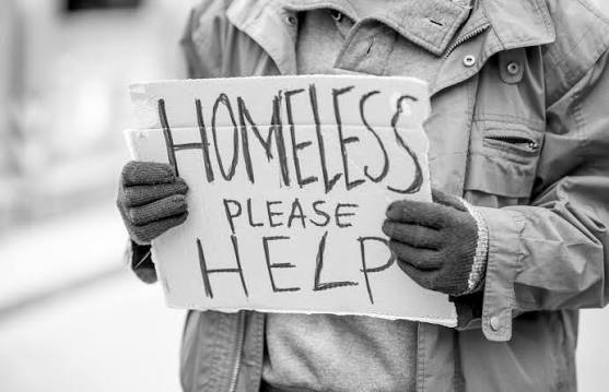 Person in a jacket holding a sign that says "Homeless Please Help" in black marker.