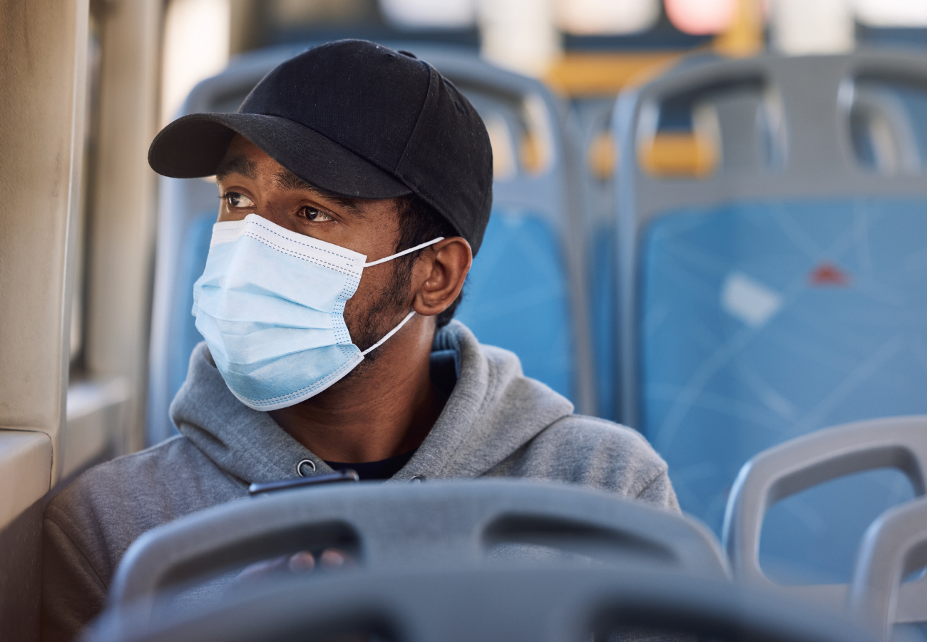 A man wearing a black baseball cap and a light blue face mask sitting on a bus, looking out the window.
