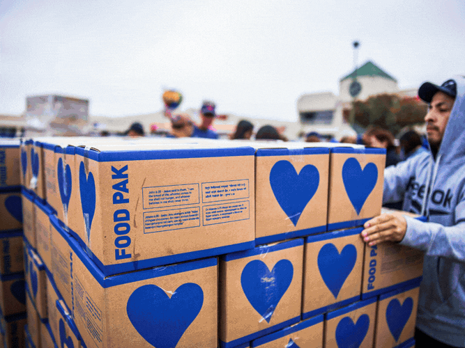A group of people, some wearing hoodies, are stacking and handling cardboard boxes labeled 'Food Pak' with a large blue heart symbol on them, outdoors.