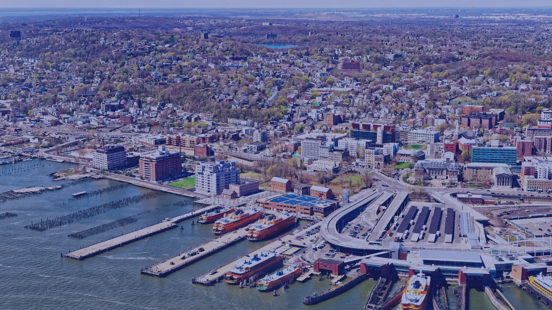Aerial view of a city with a harbor, featuring multiple boats, docks, and high-rise buildings, surrounded by a mix of urban and green areas.