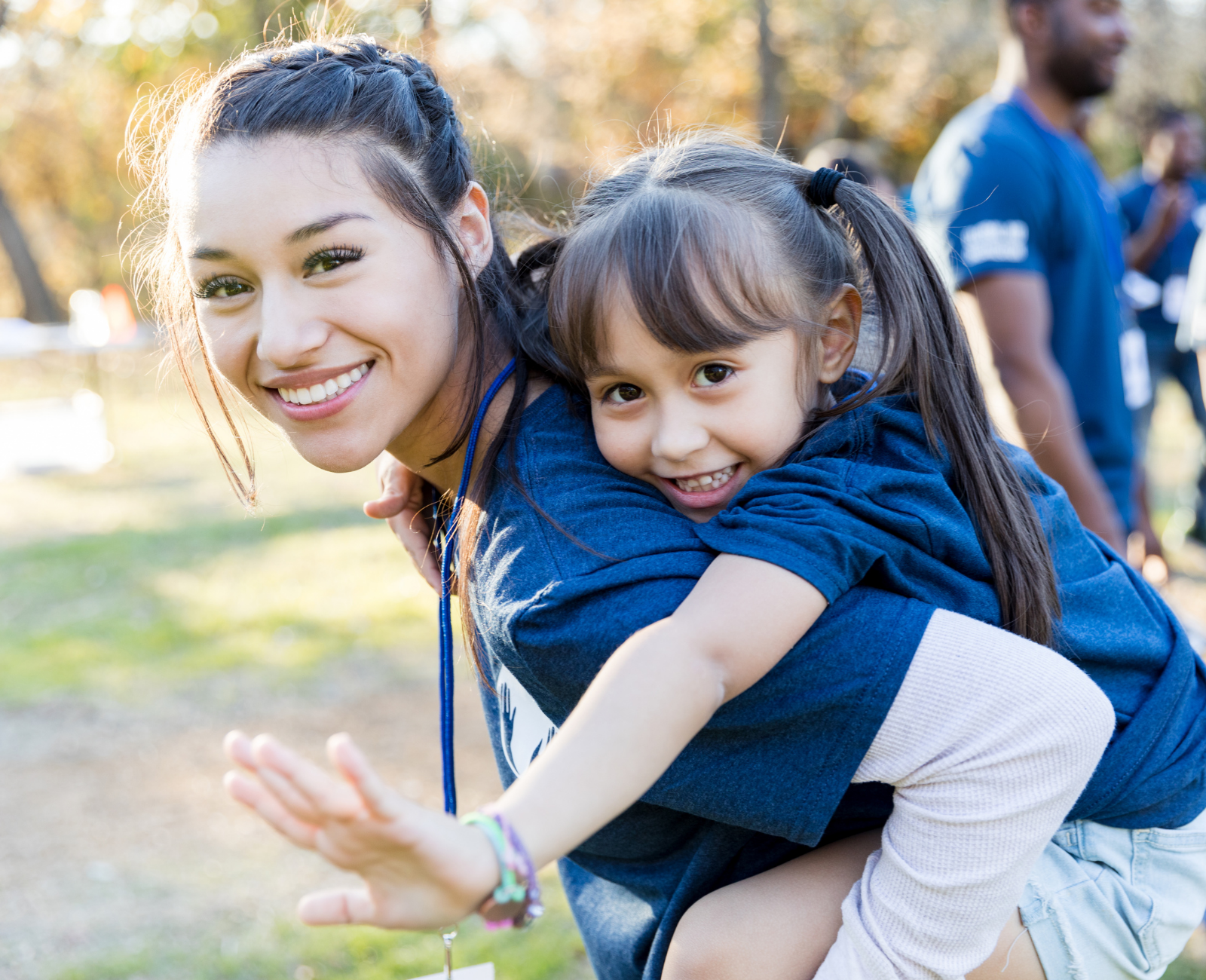 A young woman with dark hair in a braid and a young girl with dark hair in pigtails, both smiling, outdoors in a park. The woman is carrying the girl on her back, and they are both wearing blue shirts. The background has autumn-colored trees and other people, slightly blurred.