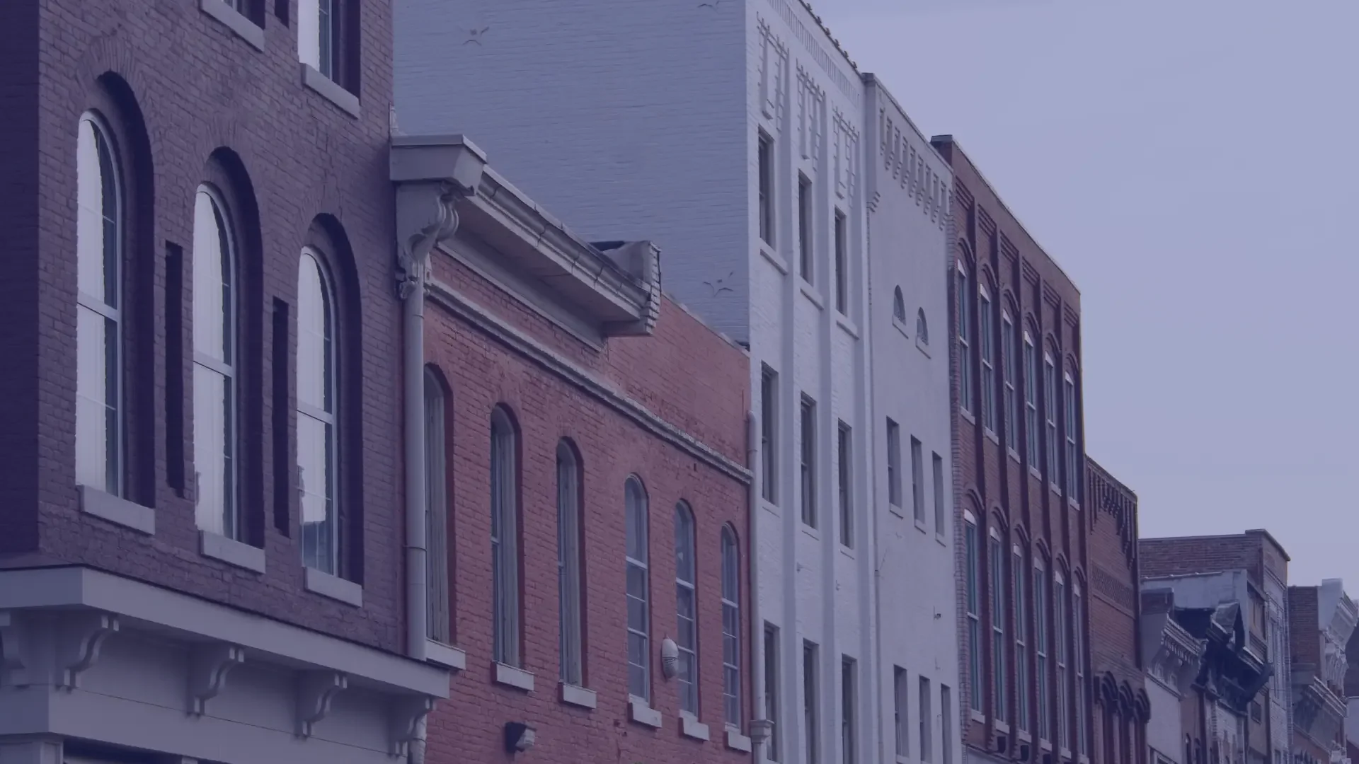 A row of multi-story brick buildings with arched windows, some with decorative moldings, on a city street during dusk or early evening.