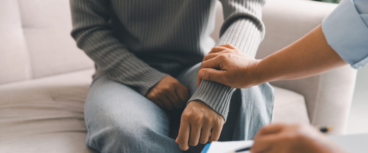 Person sitting on a couch with a hand on their wrist while another person touches their arm, suggesting a comforting or supportive interaction.