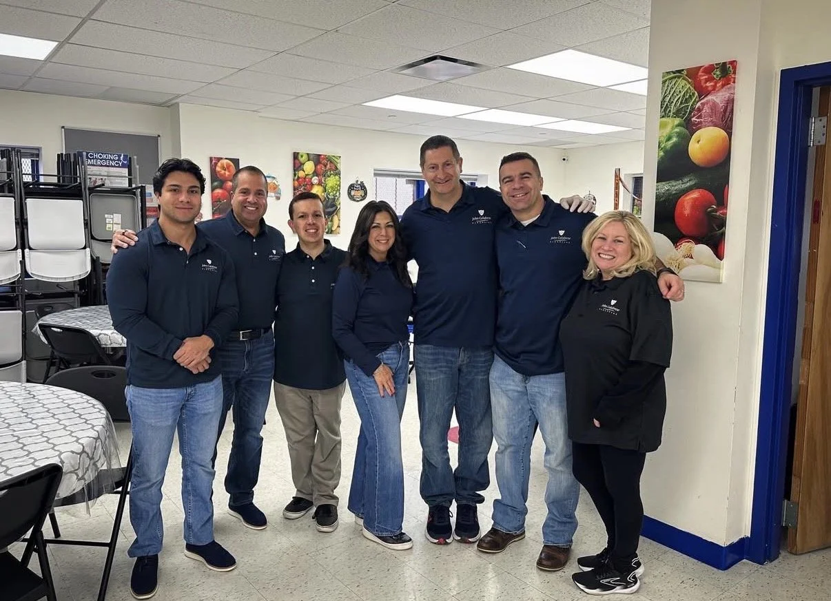 Seven smiling people standing together in a room, posing for a photo, with some wearing matching navy blue shirts.