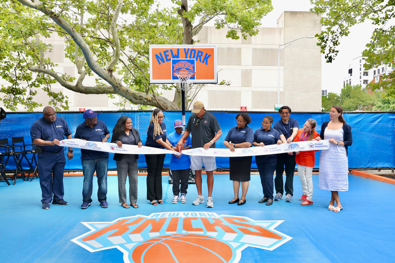 A ribbon-cutting ceremony on a New York Knicks themed basketball court, with several people standing in a row, holding a ribbon, in front of a basketball hoop and a New York Knicks logo on the court.