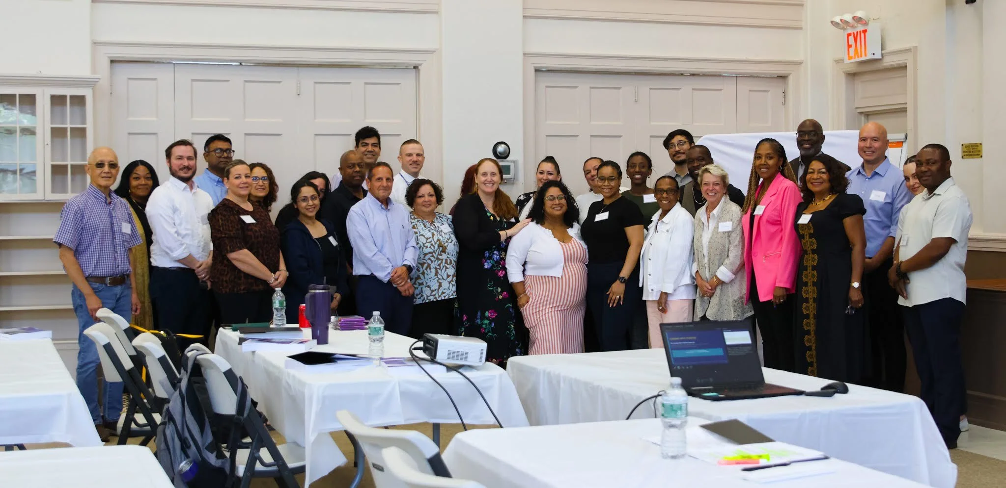 A diverse group of professionals posing together at a conference or meeting in a large room with white walls and tables.