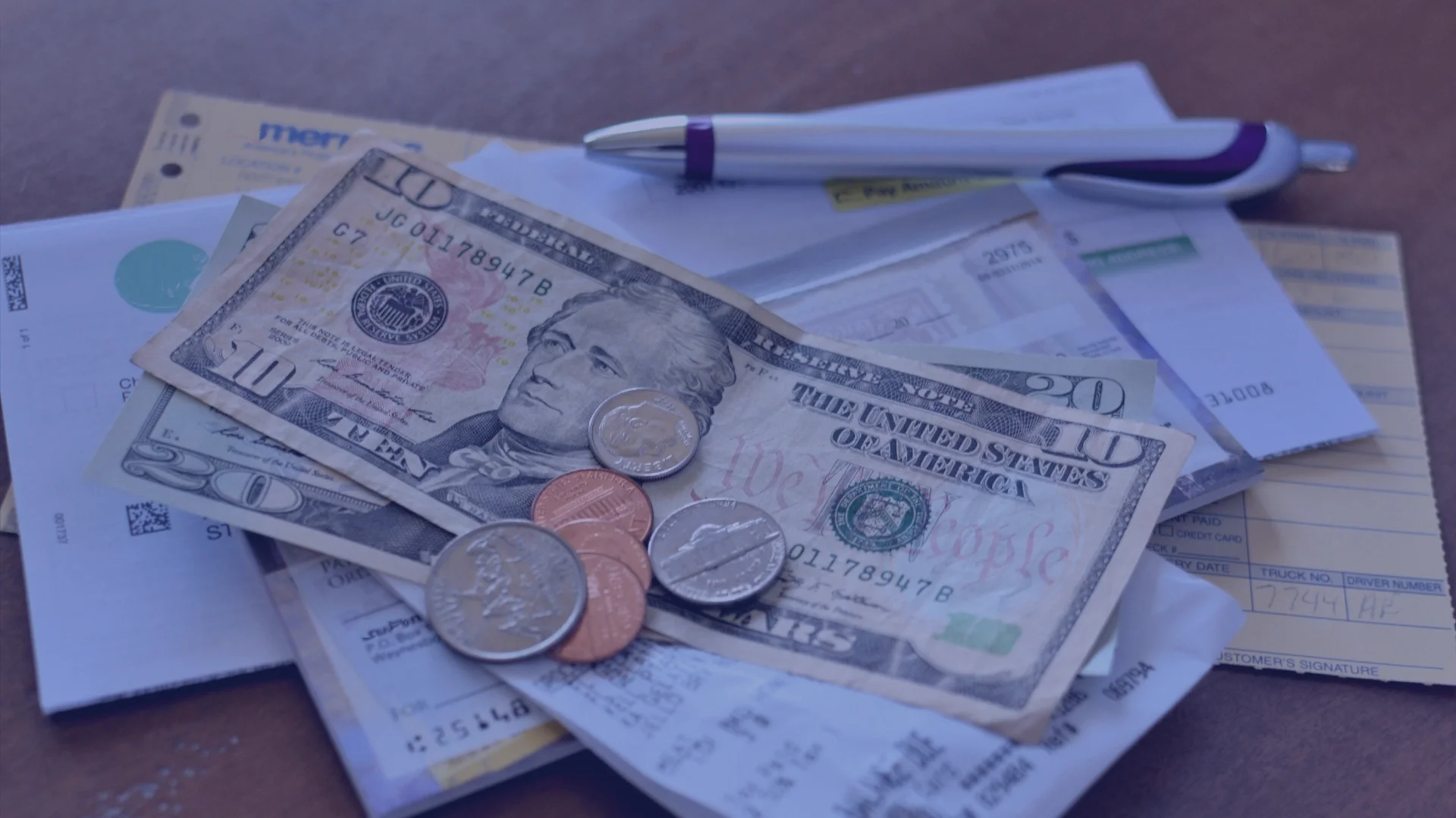 A pile of U.S. currency and receipts on a brown surface. The money includes a twenty-dollar bill, a ten-dollar bill, and several coins.