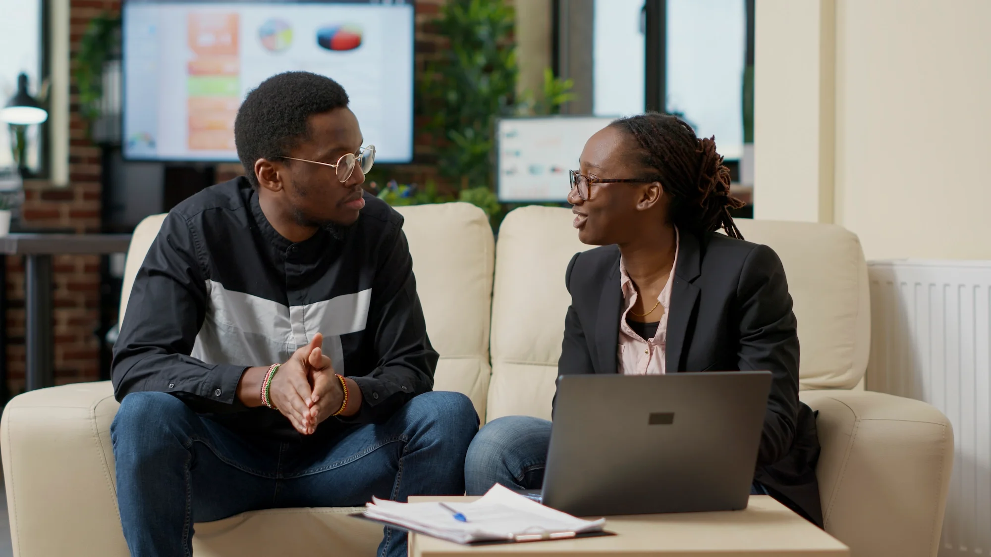A man and a woman are sitting on a cream-colored couch, engaged in a conversation. The man is wearing glasses, a black and white jacket, and has his hands clasped. The woman is also wearing glasses, dressed in a black blazer with a pink shirt underneath, and has a laptop on her lap. They are in a modern office setting with a monitor displaying colorful charts in the background.