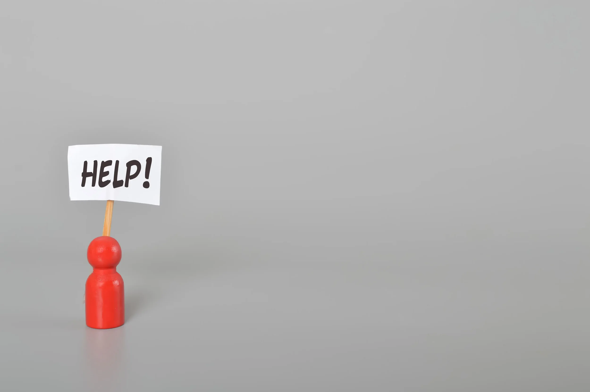 A small red figurine holding a sign that says 'HELP!' in black letters. The figurine is positioned on a flat surface with a plain gray background.