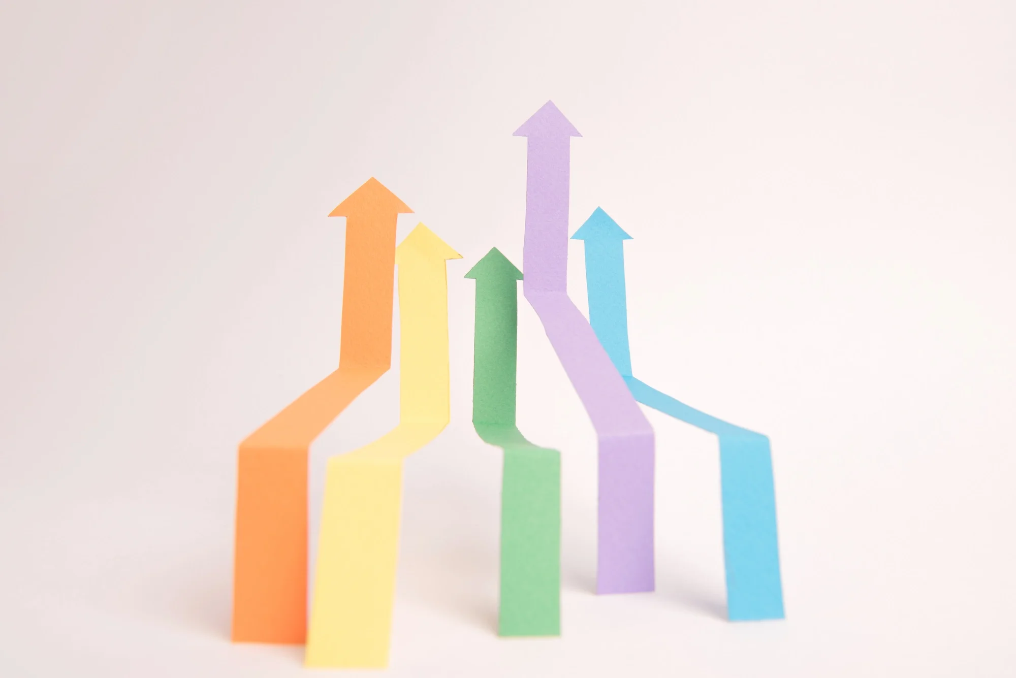 Colorful paper arrows pointing upward in orange, yellow, green, purple, and blue, arranged on white background.