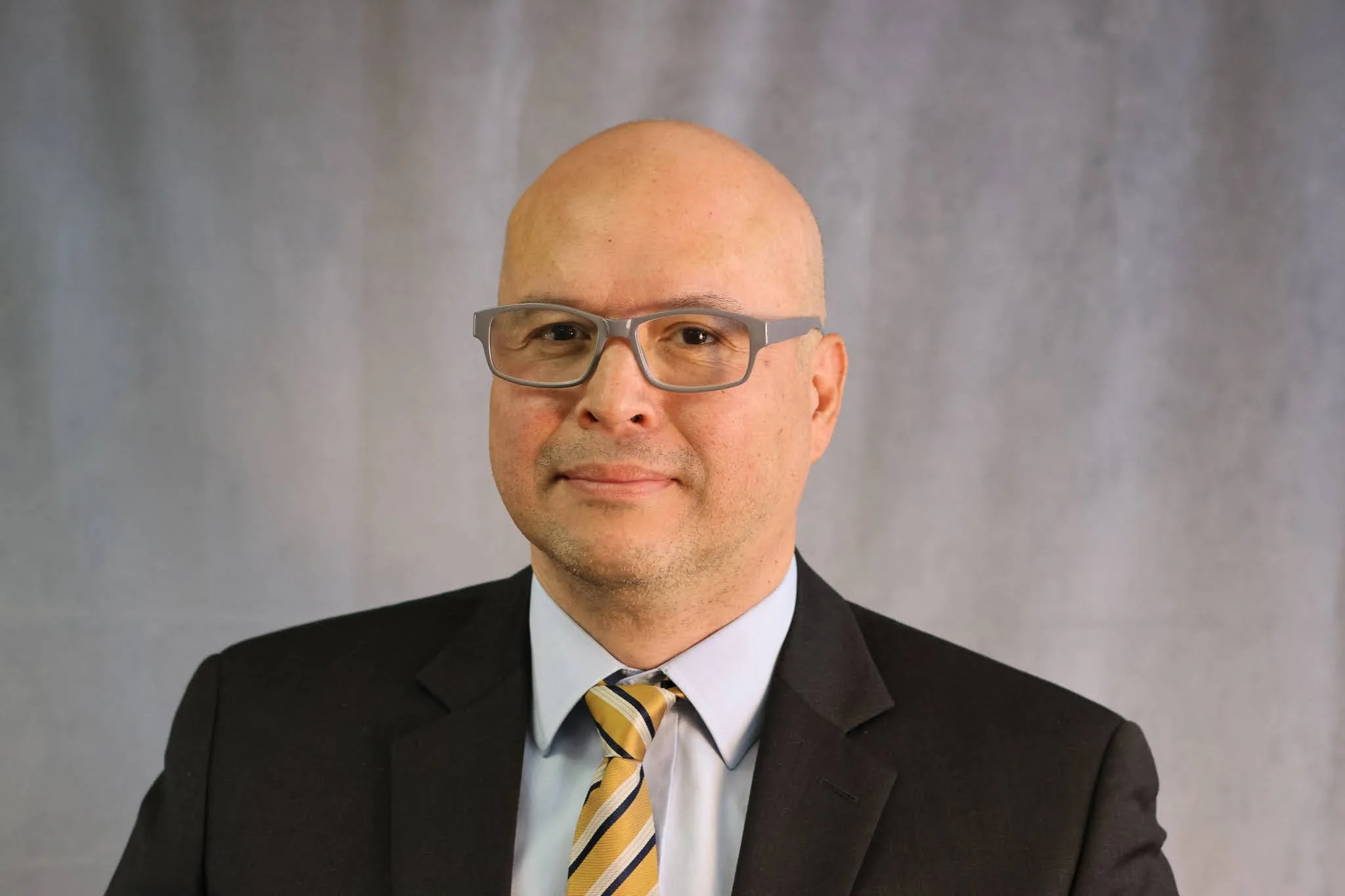 A man dressed in a black suit with a white shirt and a yellow striped tie, wearing gray glasses, posing against a plain gray background.