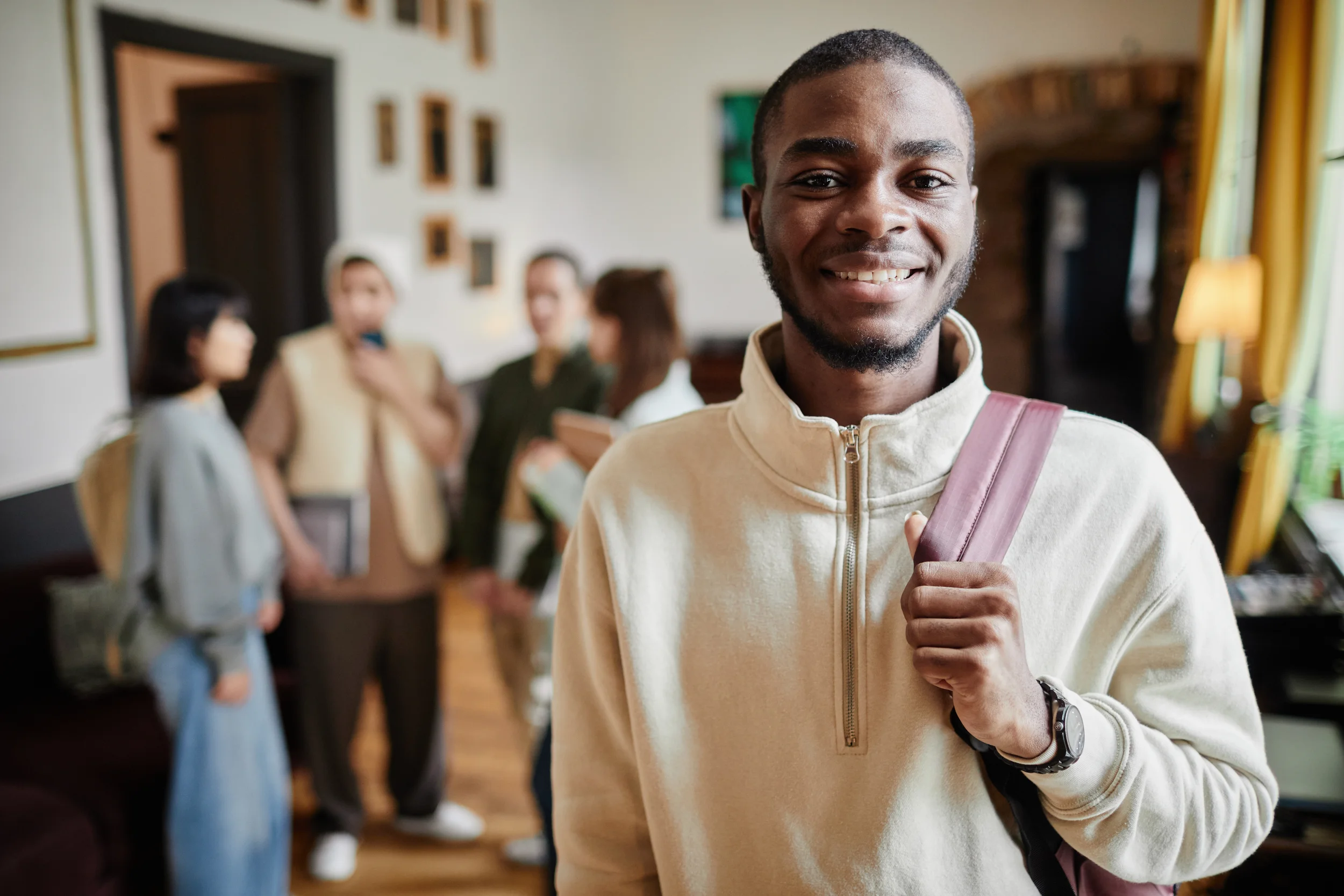 A young man with a backpack smiling at the camera in a room with several people in the background.
