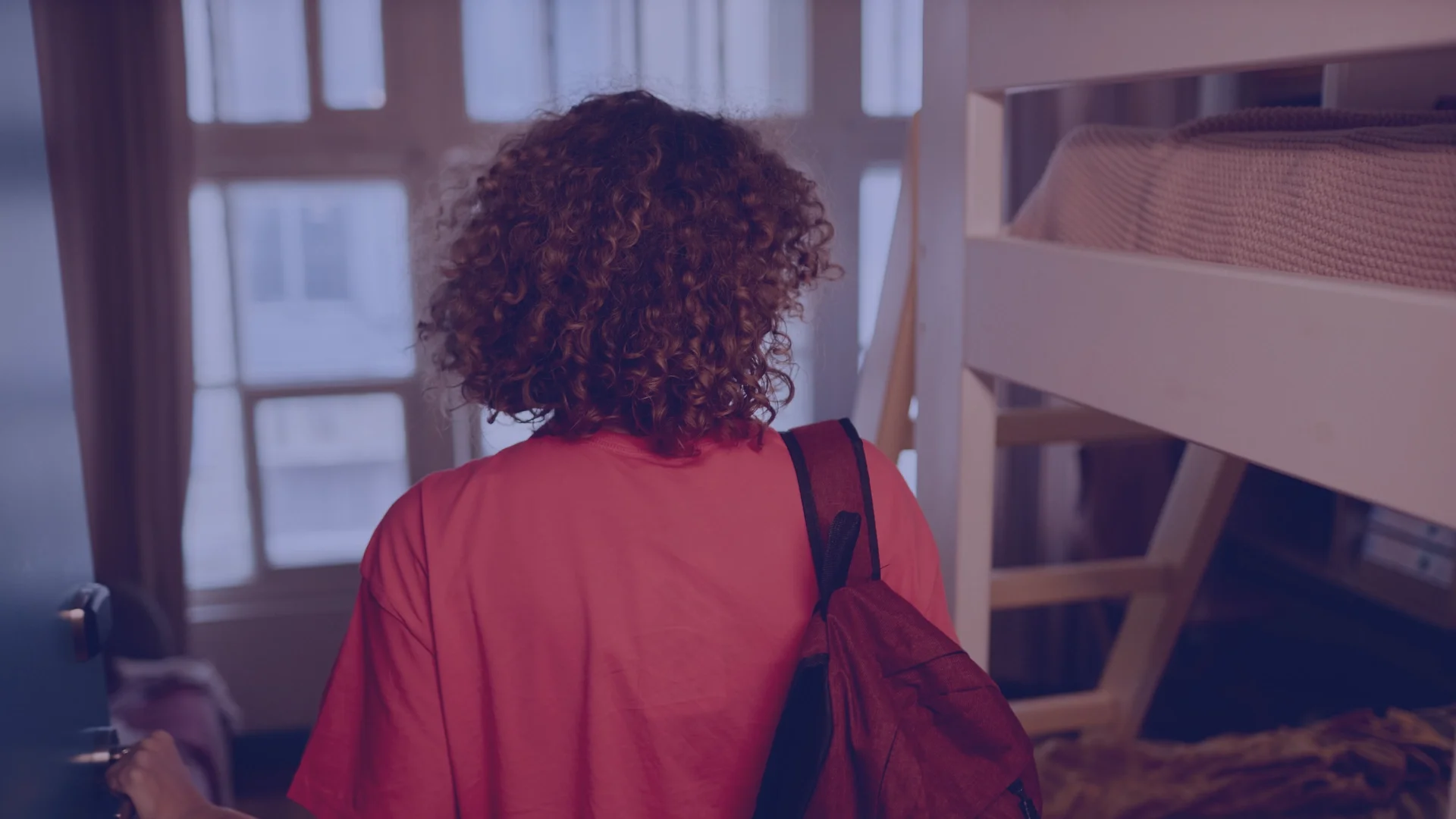 A person with curly hair wearing a red shirt and a backpack, standing inside a room with a bunk bed and a window.