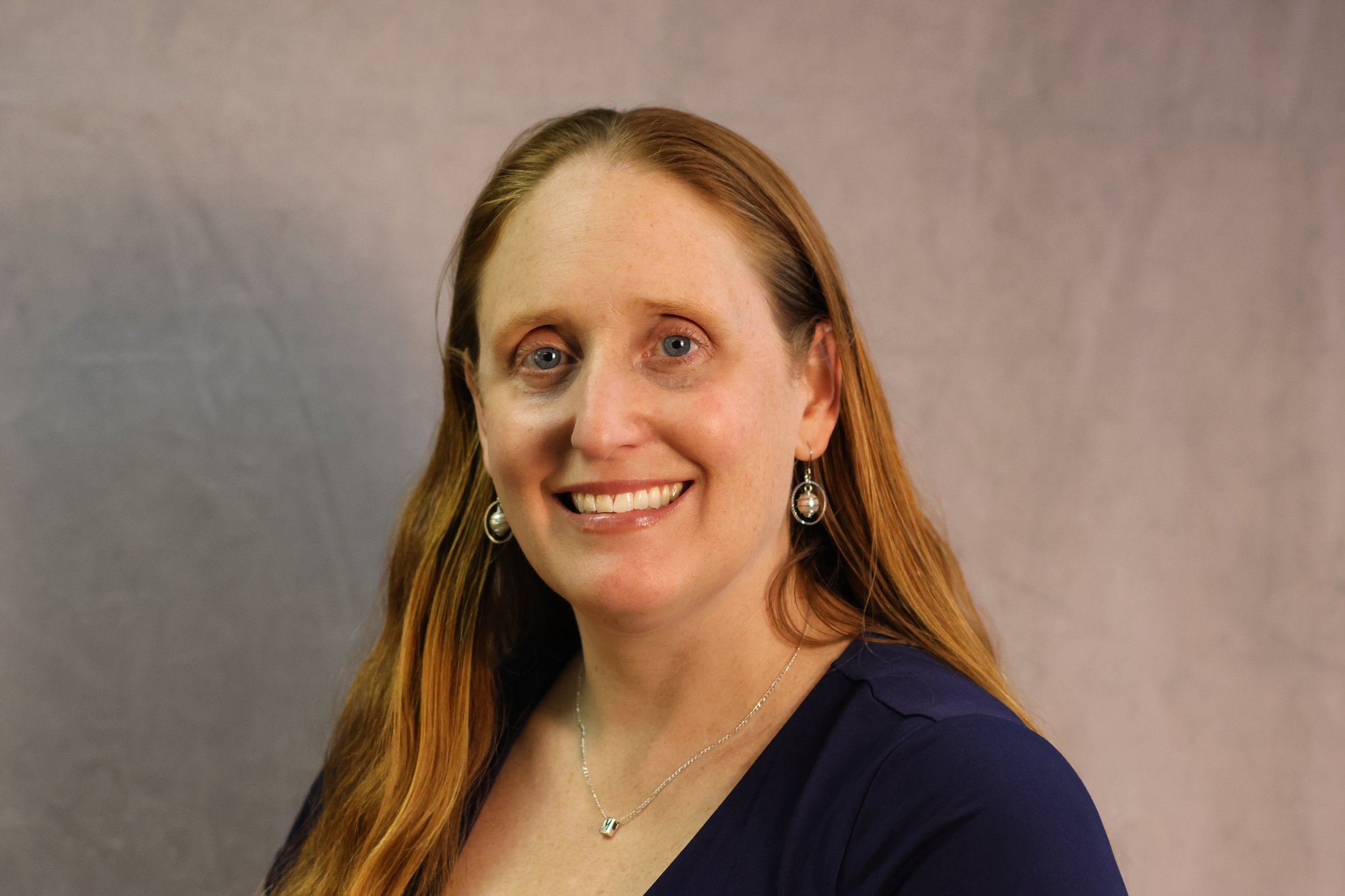 A woman with long red hair, light skin, and blue eyes smiling at the camera, wearing earrings, a necklace, and a navy blue top, standing in front of a plain gray background.
