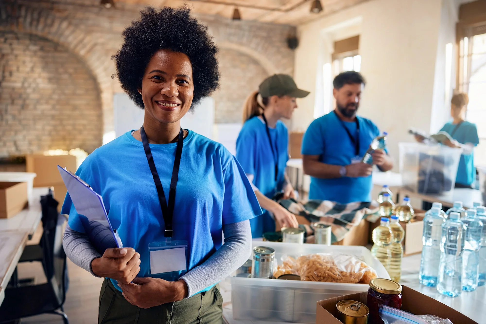 A smiling woman in a blue shirt holding a clipboard in a room with brick walls and large windows. In the background, three people are working at a table with canned food and bottled water, wearing similar blue shirts.
