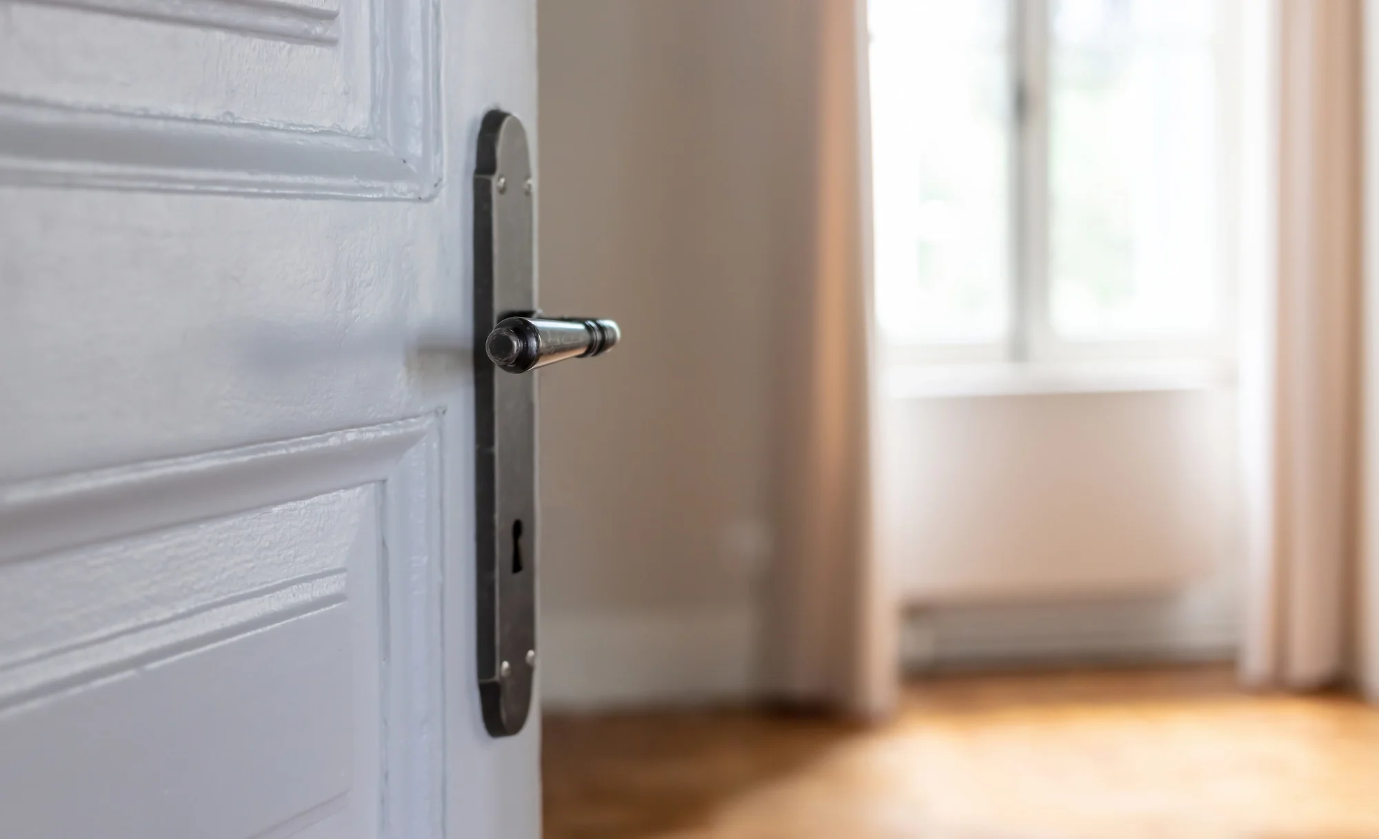 Close-up of an open door with a metal latch and a black handle, leading into a room with a window and beige walls.