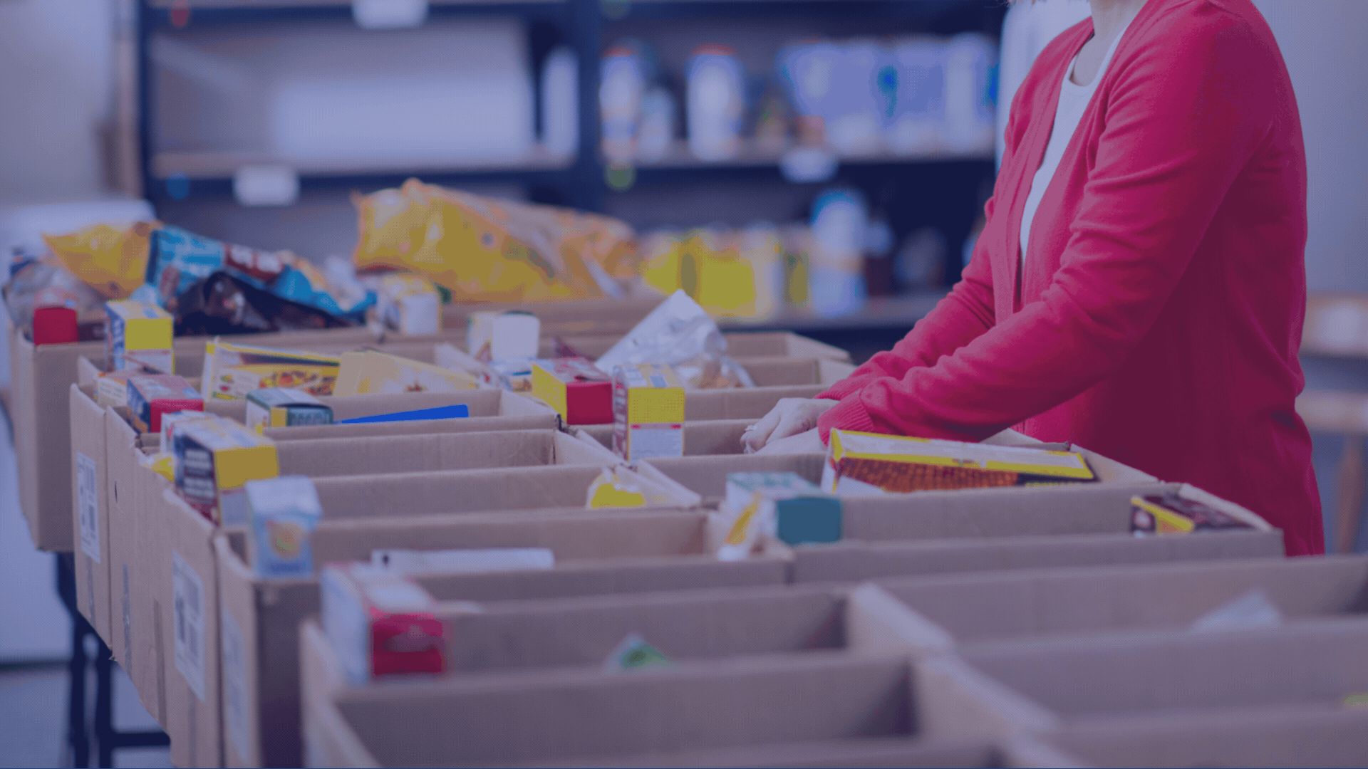 A person in a red cardigan organizing or sorting items in a large cardboard box filled with various packages or products in a storage or warehouse setting.