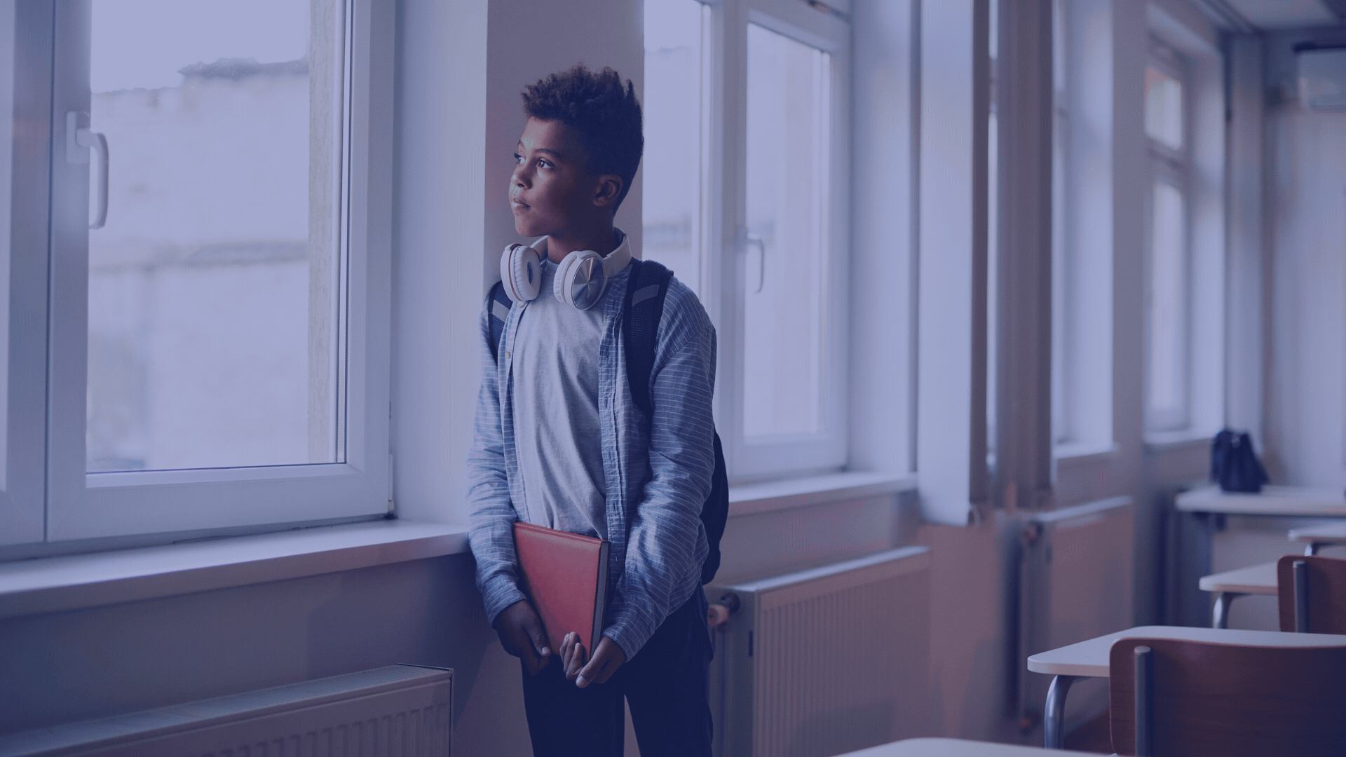 A young boy stands in a classroom near a large window, wearing headphones around his neck, holding a red folder, and looking thoughtfully outside.