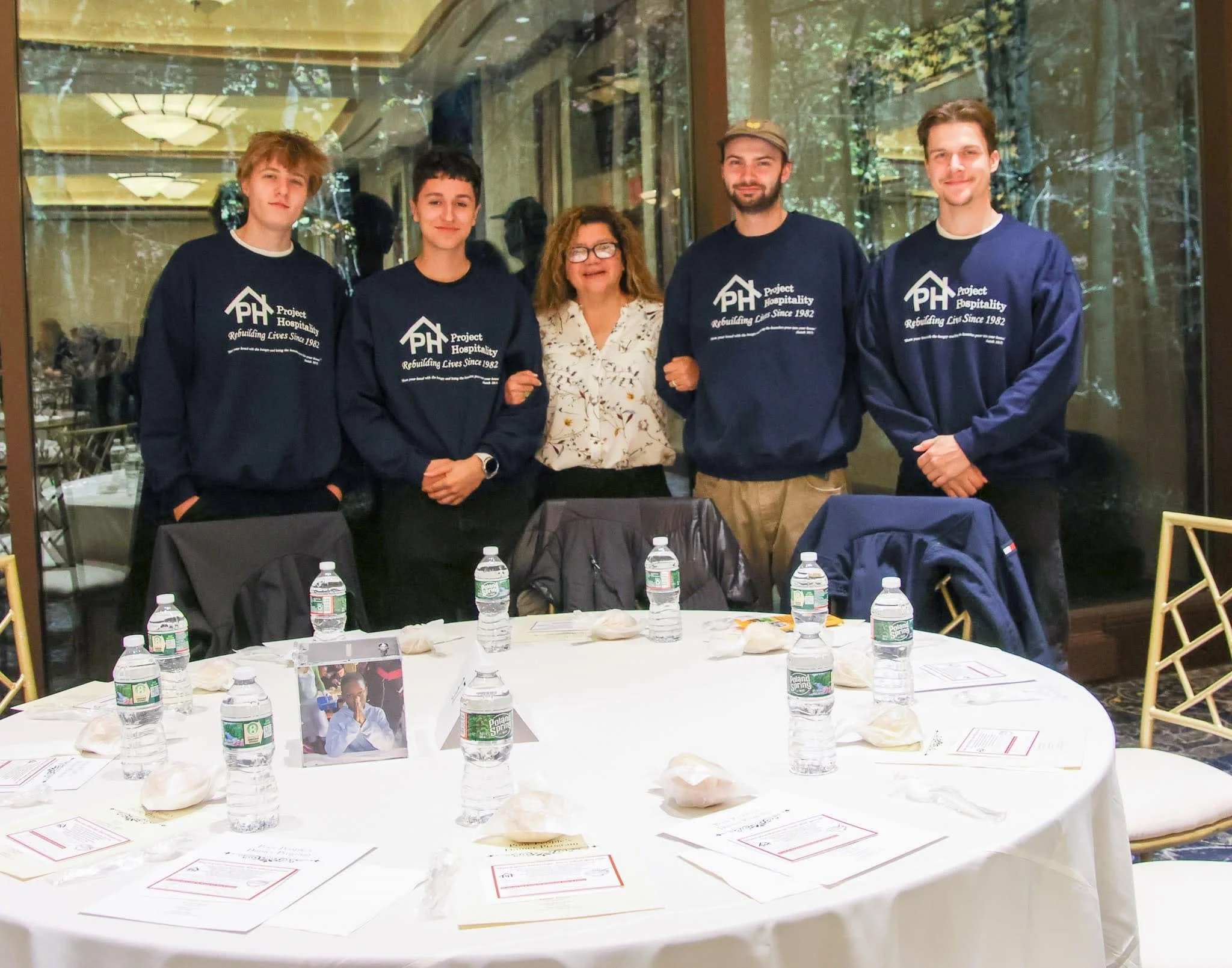 Five people standing behind a table with bottled water, pastries, and printed papers, in a well-lit indoor setting, smiling at the camera.