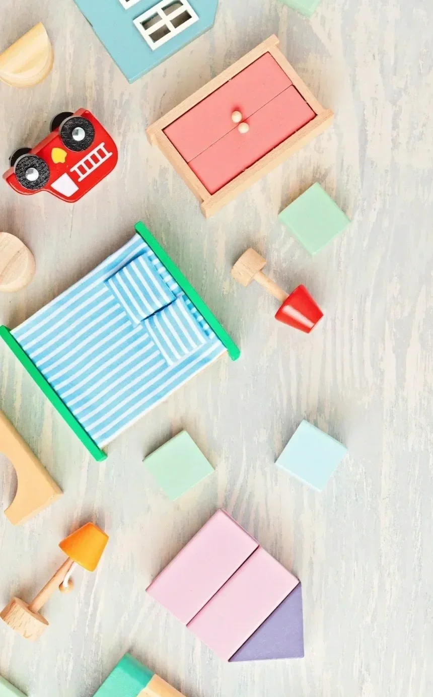Colorful wooden toy furniture and accessories scattered on a light wooden surface, including a red car-shaped toy, a blue striped bed, various blocks, and small lamps.