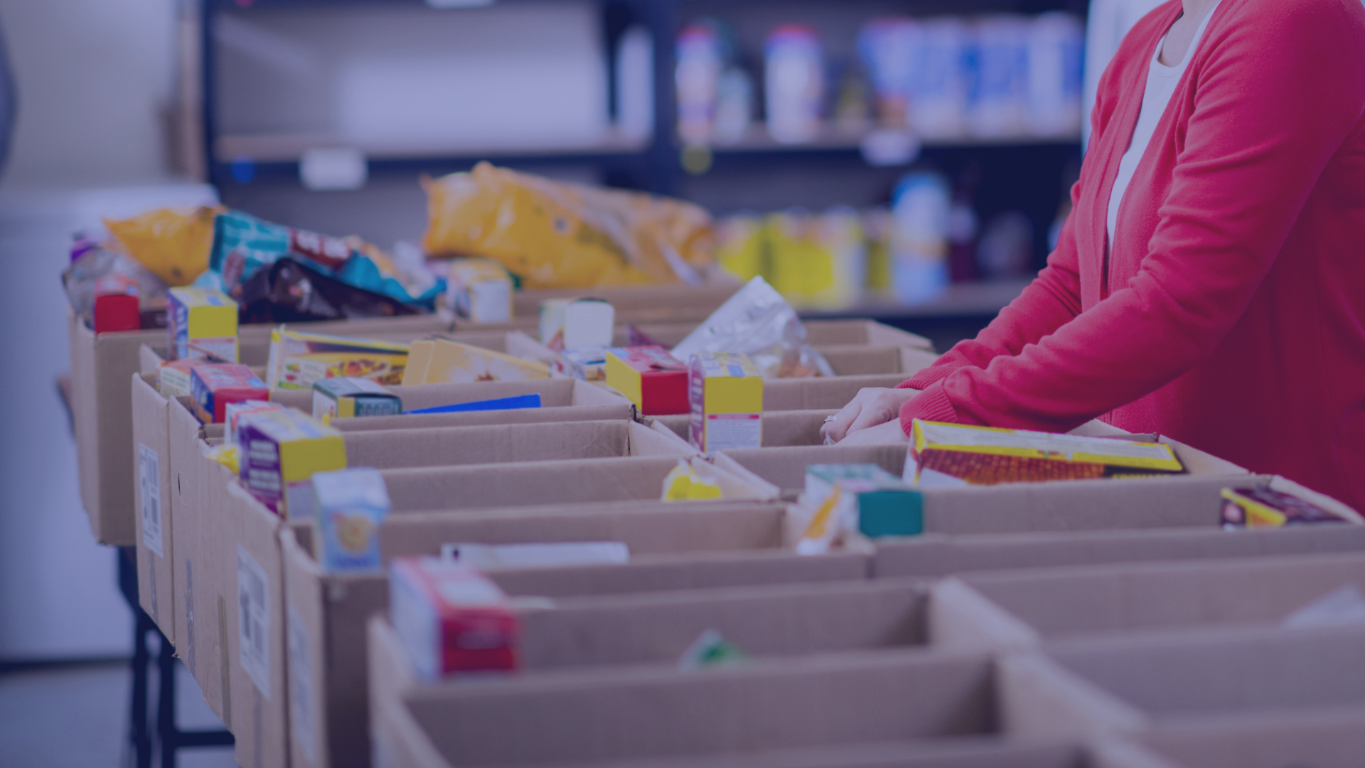 Person packing food items into cardboard boxes in a warehouse or food bank setting.