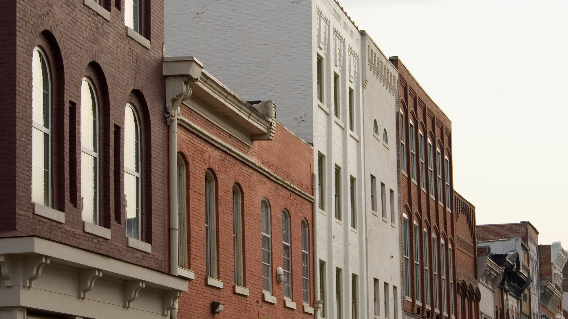 Row of multi-story brick buildings with various architectural details, including arched windows, decorative moldings, and gutter systems, under a clear sky.