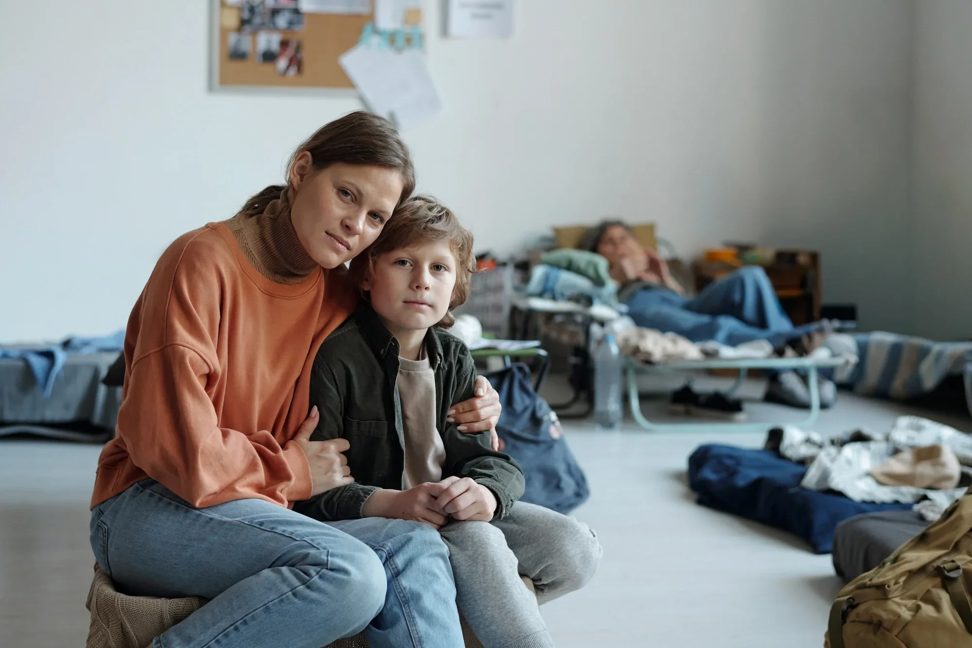 A woman with brown hair tied back wearing an orange sweater and blue jeans, sitting on the floor with a young boy who has curly blond hair, wearing a black jacket and grey pants, in what appears to be a shelter or temporary living space. They are sitting close together, with the woman holding the boy close. The background shows a notice board, scattered bags, and belongings on the floor.