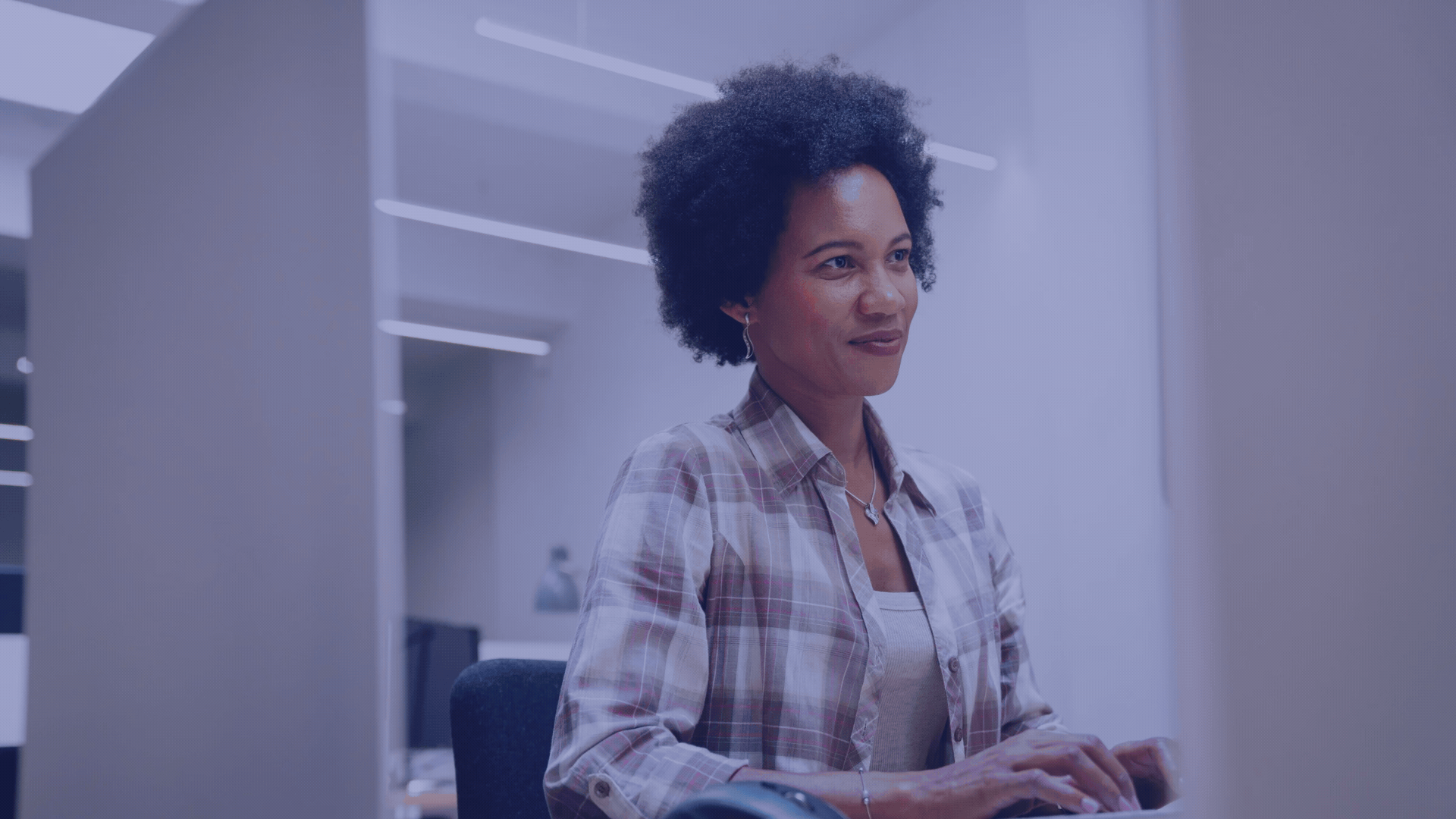 A woman with short curly hair wearing a plaid shirt sitting at a desk in an office environment