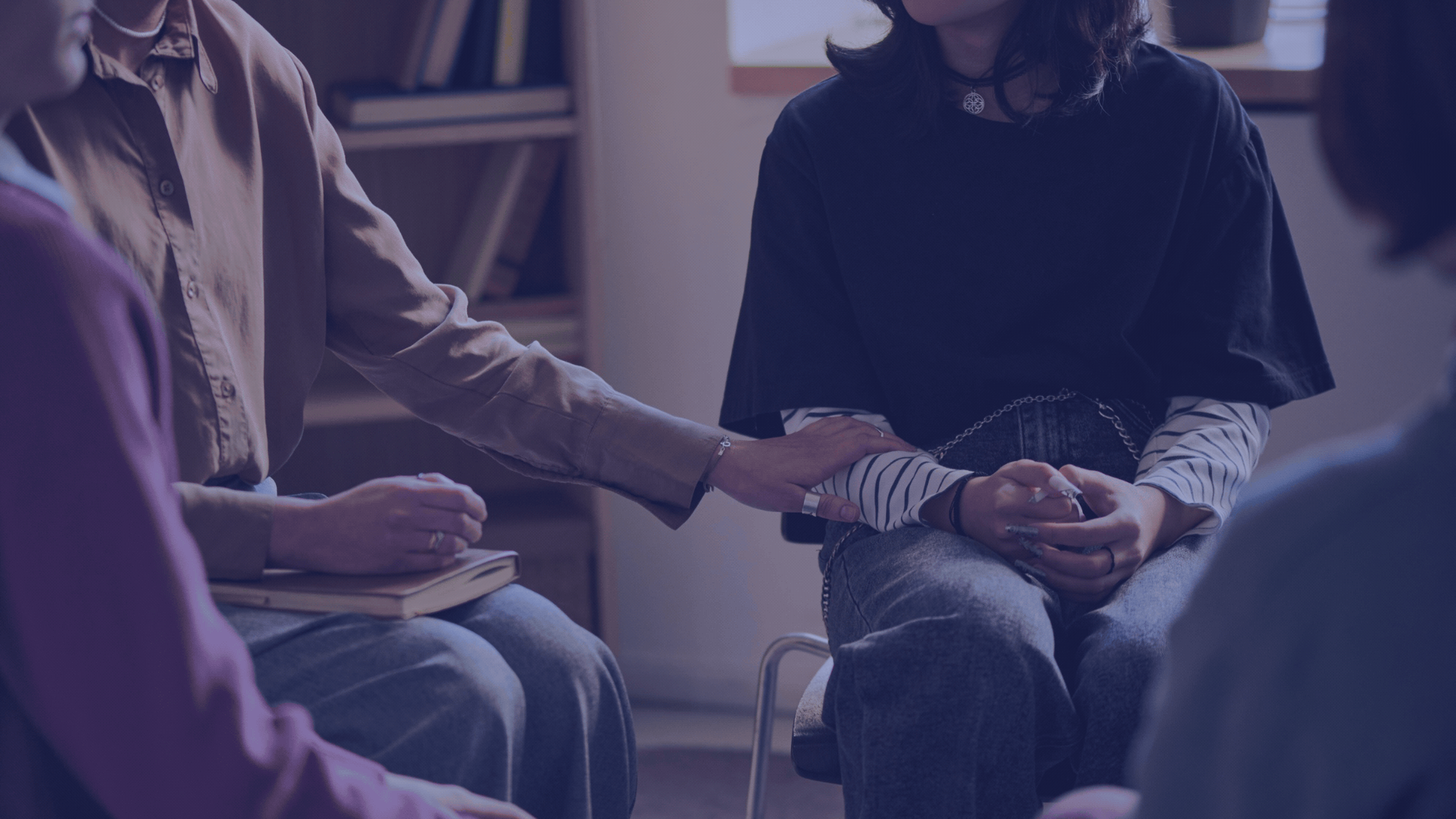 A group of people sitting together in a circle, with one person reaching out and touching another person's arm in a supportive gesture.