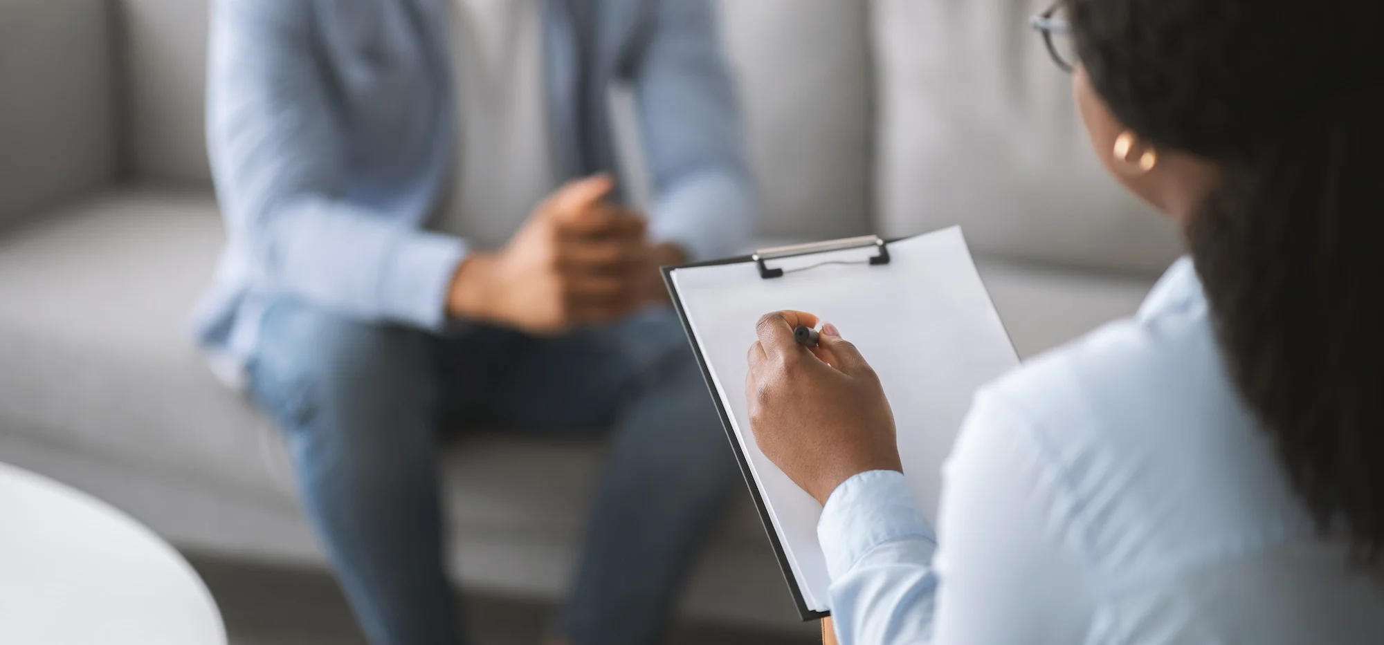 A woman with dark hair and glasses taking notes on a clipboard during a therapy session with a person sitting across from her.