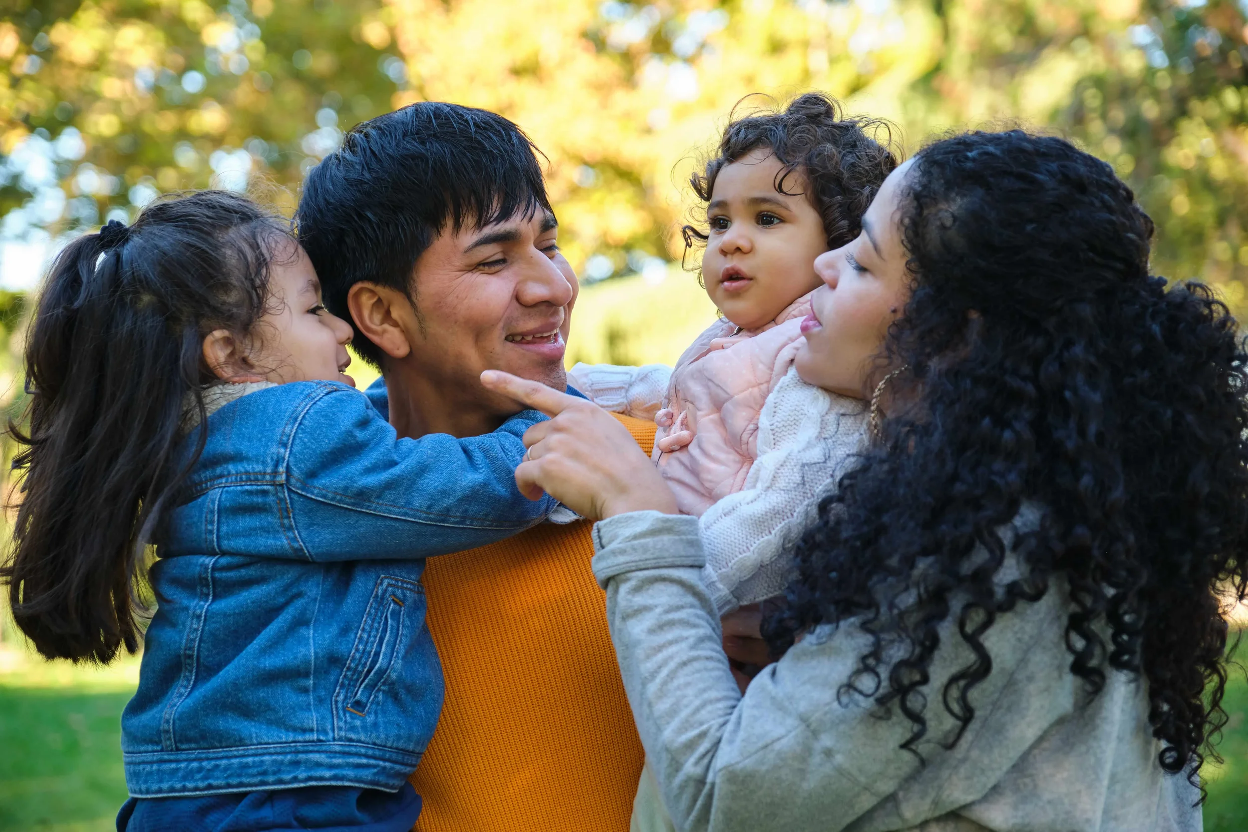 A group of four people, including two women and two children, are outdoors in a park with trees in the background. They are smiling and interacting closely, with one woman holding a young girl and the other woman touching the child's chin.