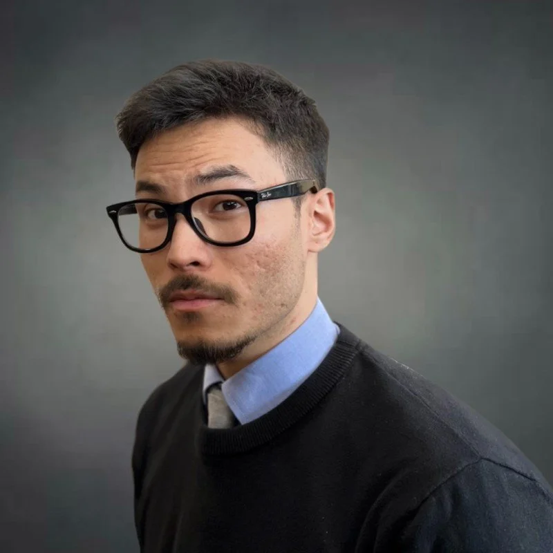 A young man with glasses, short dark hair, and facial hair, wearing a blue collared shirt under a black sweater, posing against a plain dark gray background.