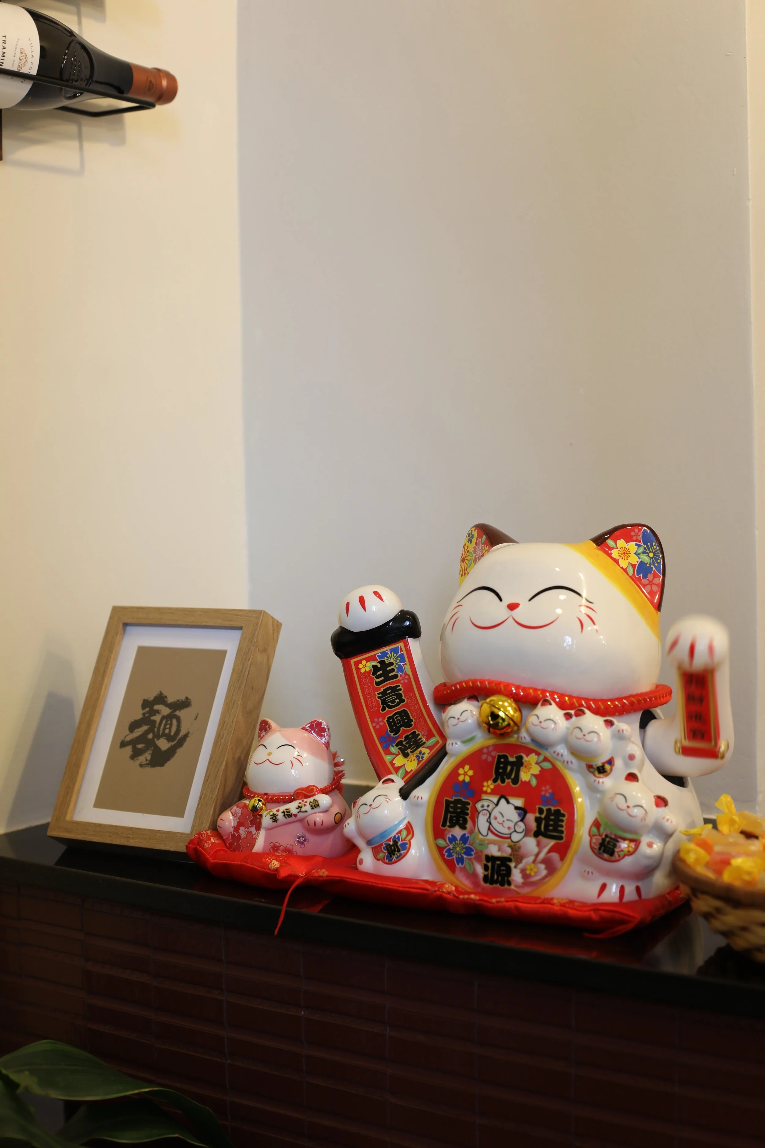 Statua di gatti Maneki-neko di diversi colori e dimensioni con simboli cinesi, presenti su un mobile con un quadro con un carattere cinese in cornice di legno.