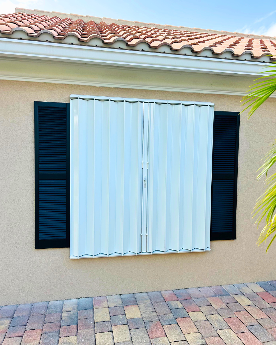 Exterior wall of a house with a white folding security door, black shutters, beige stucco wall, terracotta roof tiles, and a multicolored brick paver walkway.