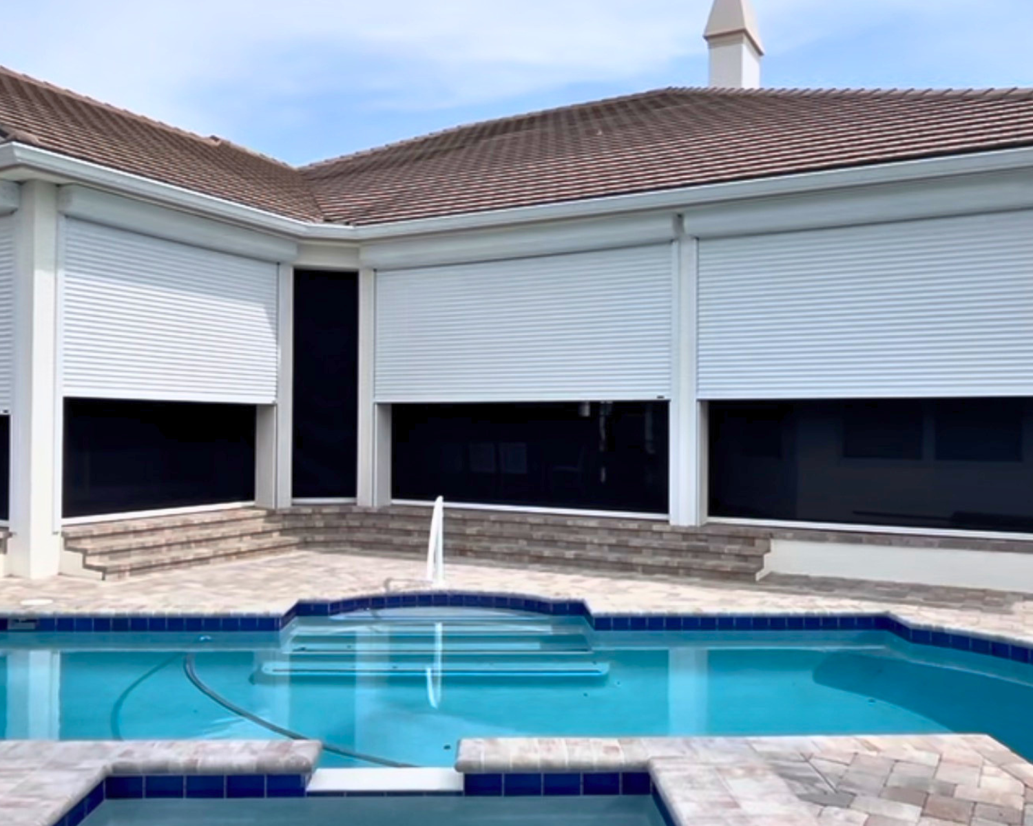 Backyard swimming pool with steps, surrounded by a stone patio, with a house in the background featuring large windows with closed white blinds and a brown tiled roof.