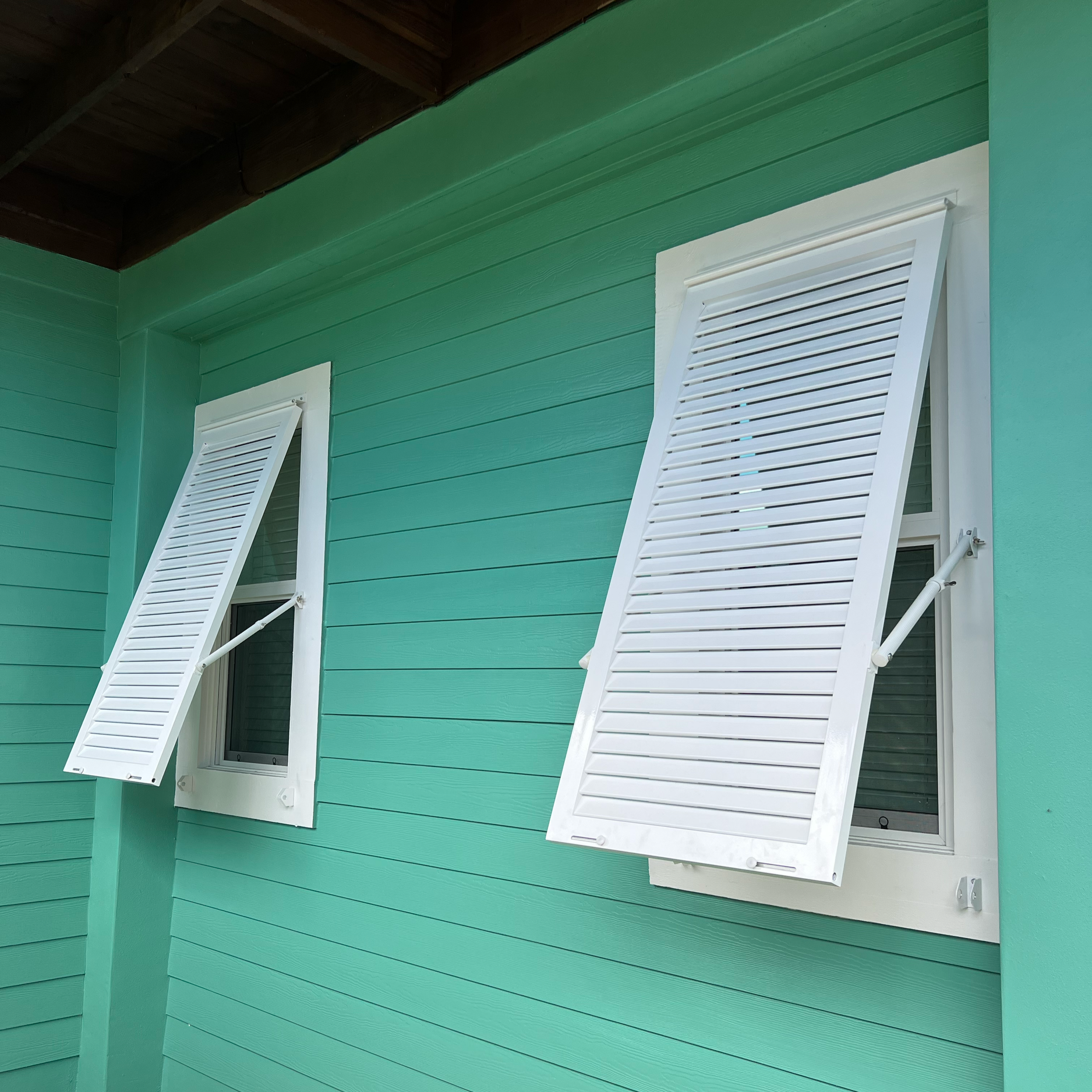 Close-up of a house's exterior showing two white window shutters on a teal-colored wooden wall, with a dark brown wooden roof above.