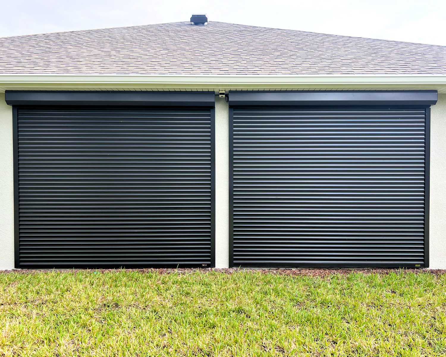 Two black metal garage doors with horizontal slats, set against a cream-colored exterior wall, with a brown shingled roof above and a green lawn in front.