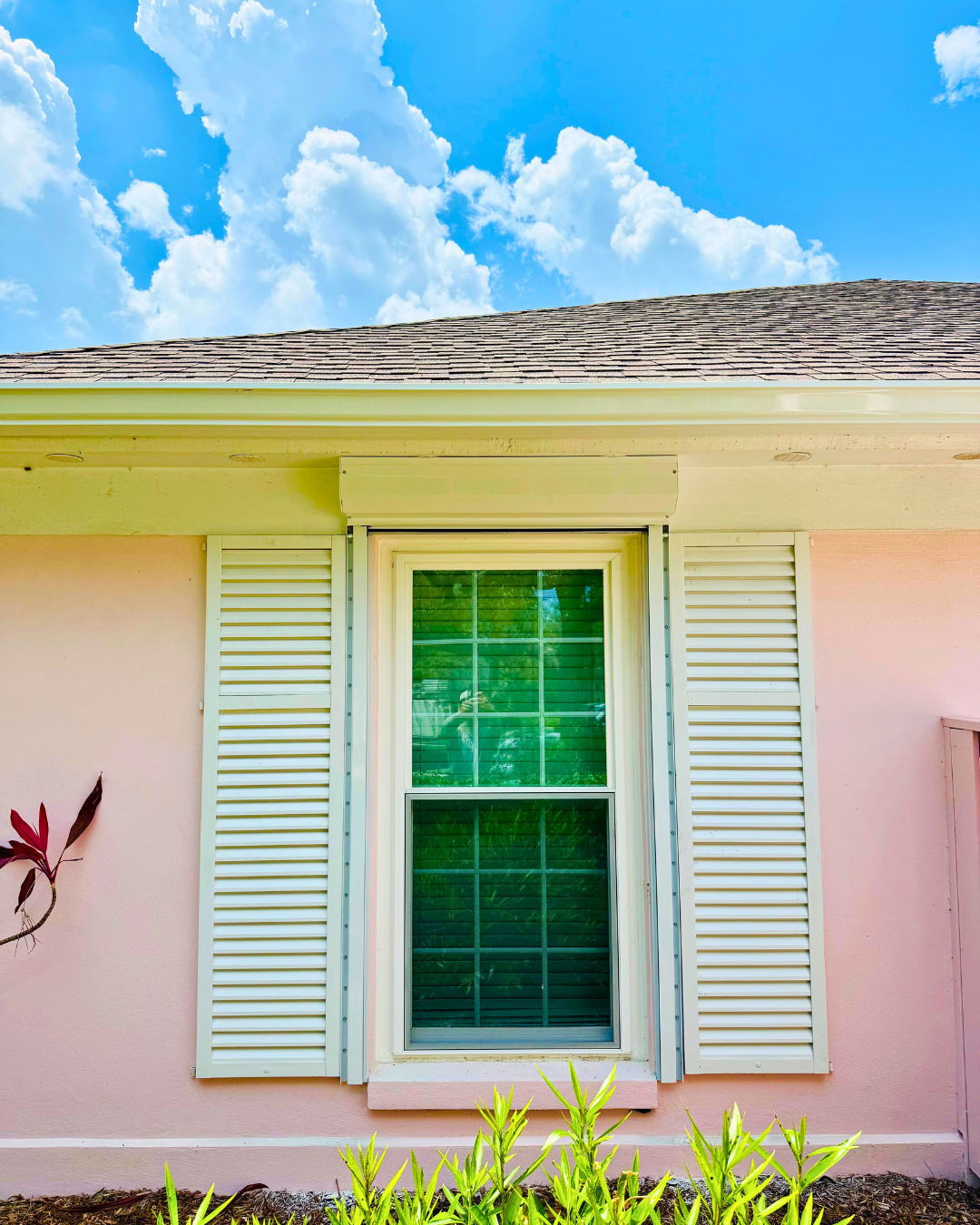 Close-up of a house window with white shutters, pink exterior wall, and a brown shingled roof, under a blue sky with white clouds.
