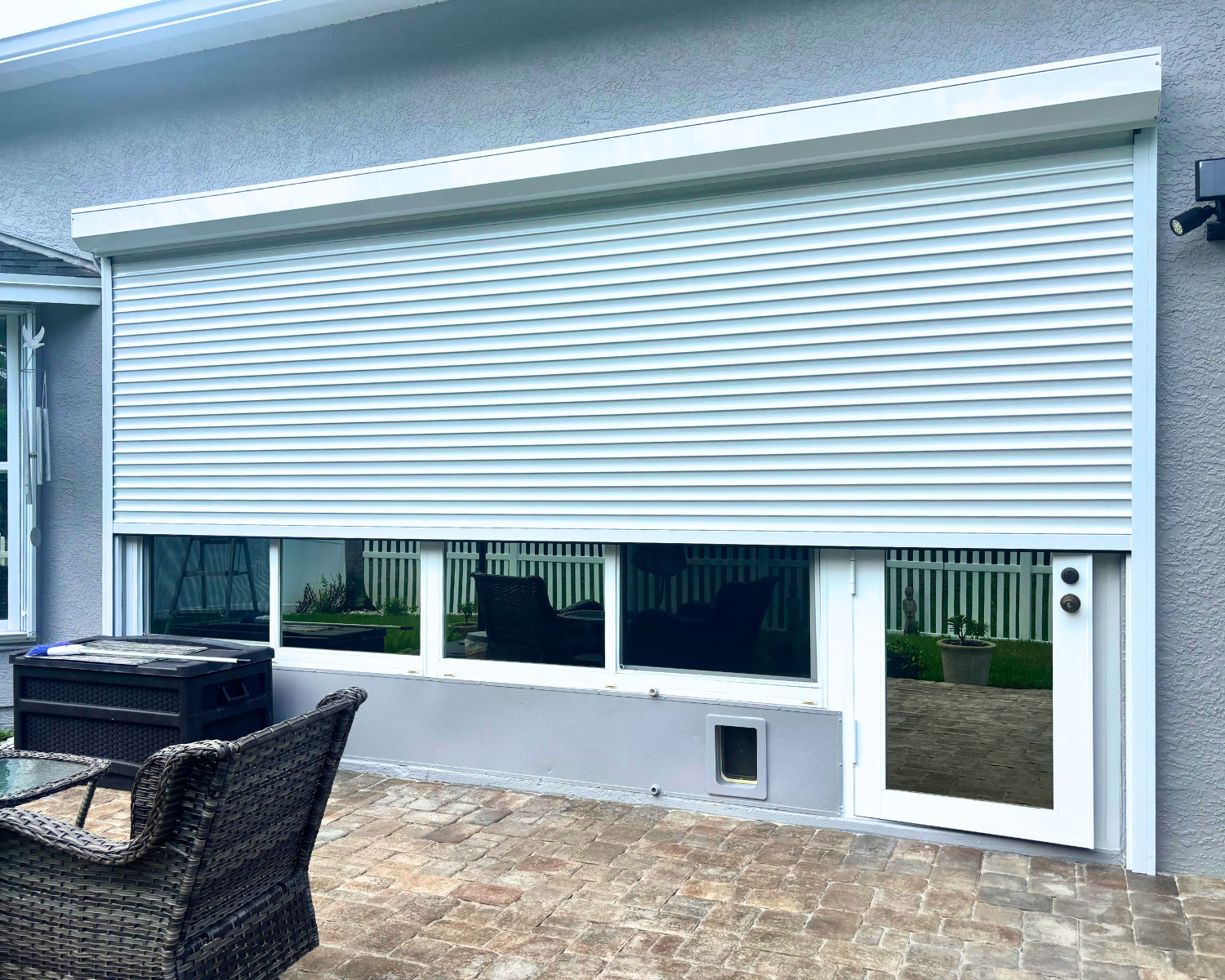 A patio area with wicker chairs and a black storage box, showing a house exterior with a large window and a sliding glass door covered by white closed rolling shutters.