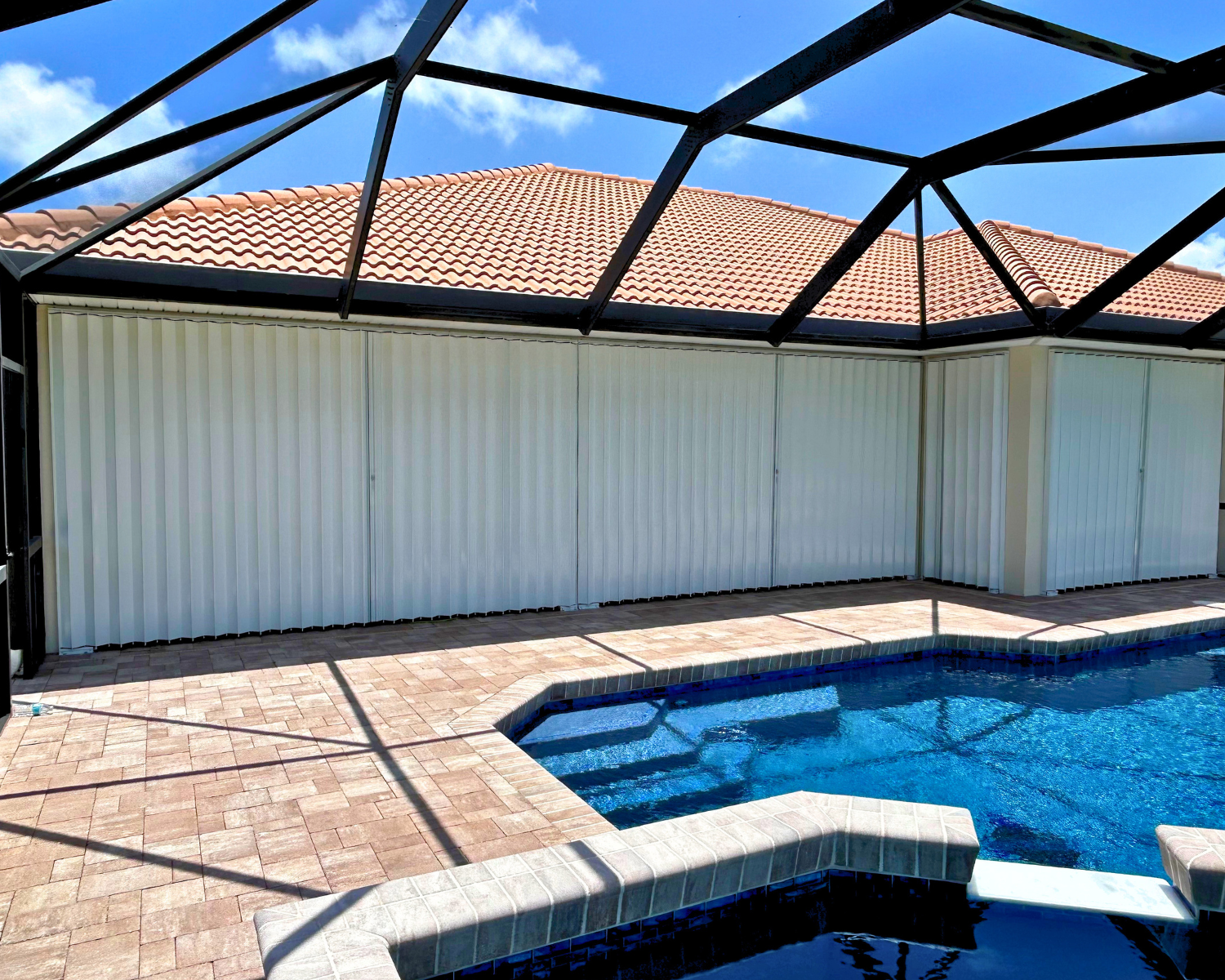 Un screened-in backyard patio area with a pool, brick pavers, a white privacy wall, and a roof with red tiles.