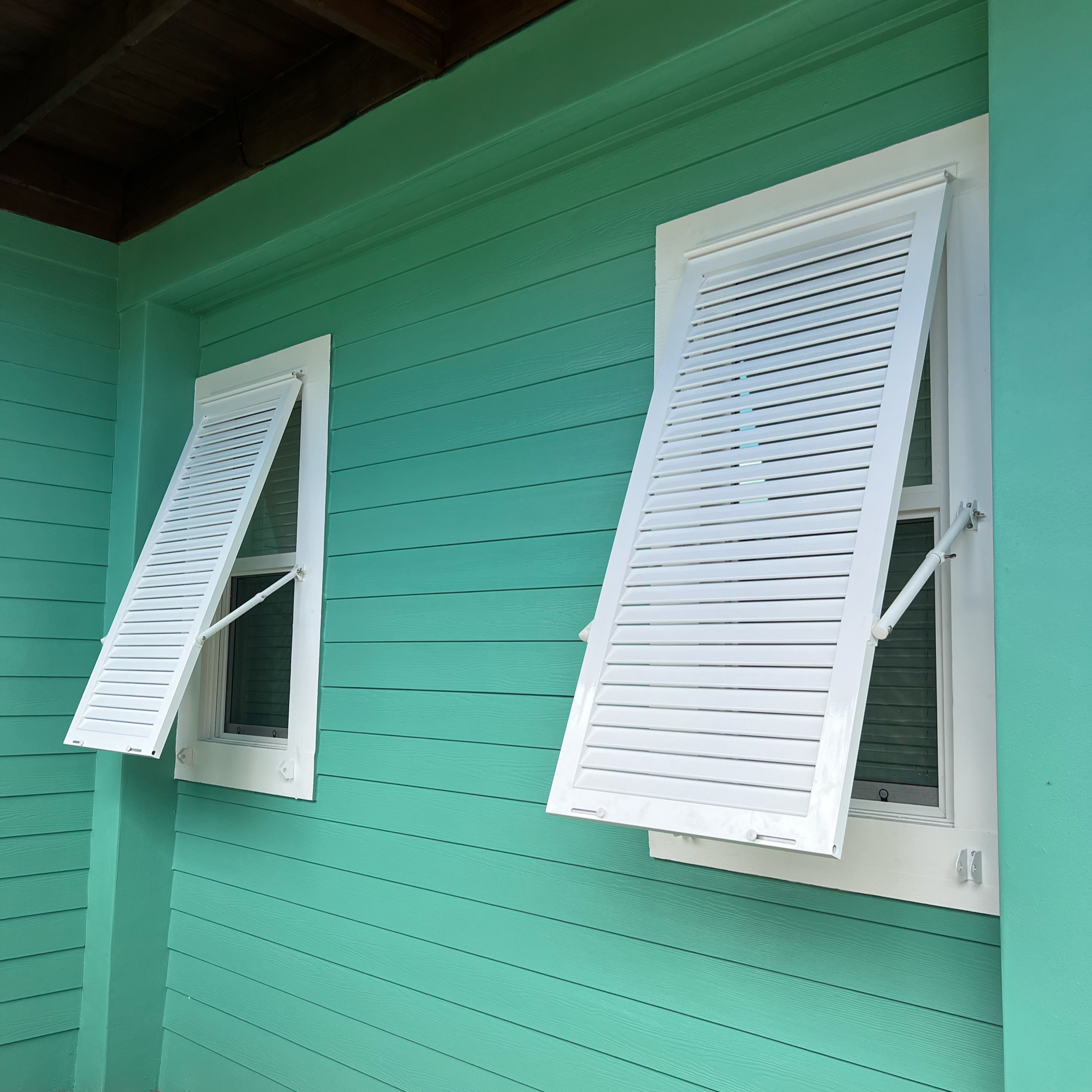 A teal house with two white framed windows, one partially open with white shutters, on a porch with a wooden roof.