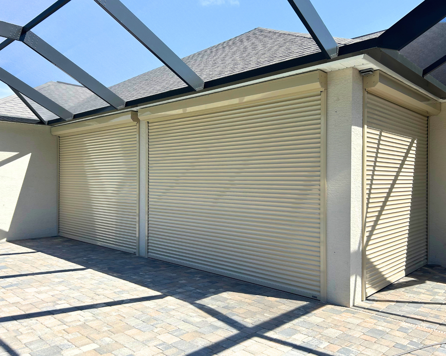 Three beige garage doors with metal roll-up shutters on a house, under a screened porch roof with a paved stone driveway.
