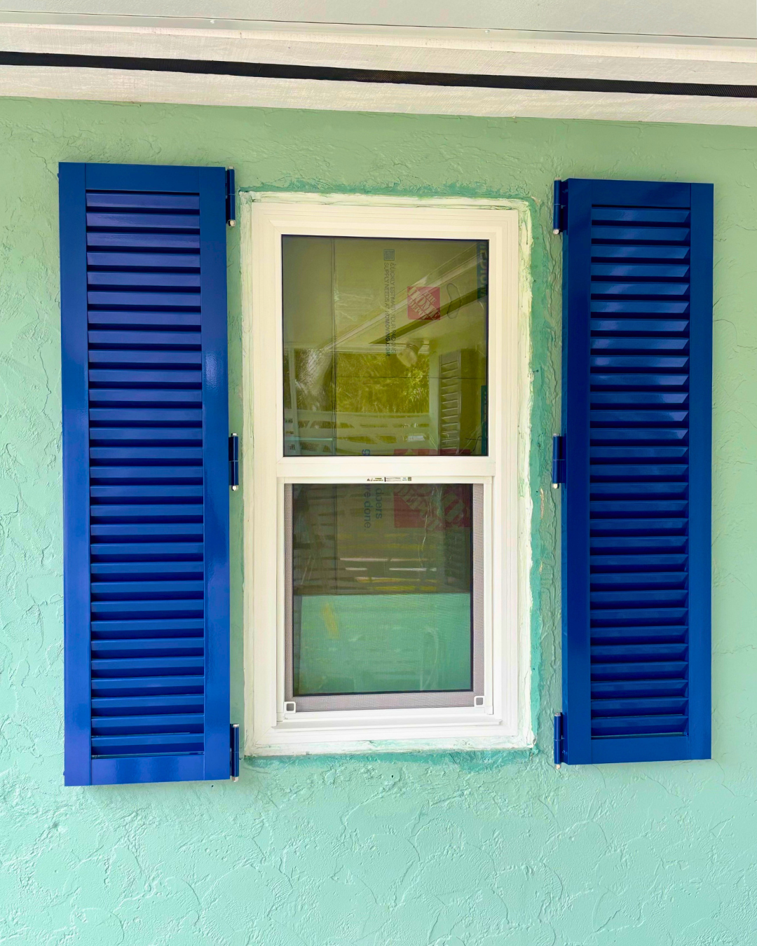 A white window with blue shutters on a mint green stucco wall.