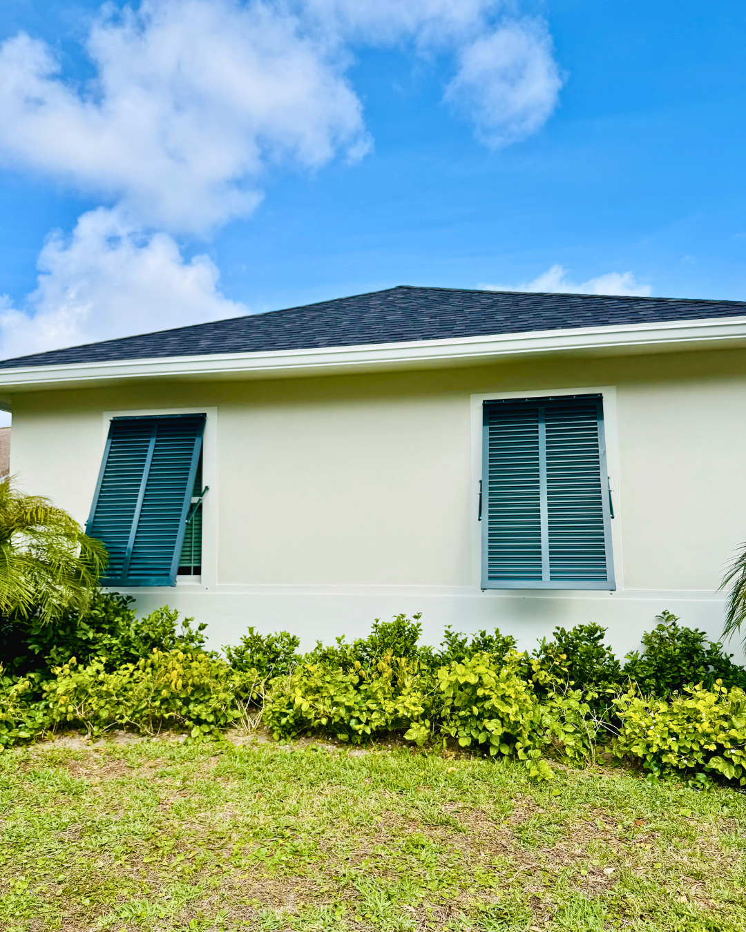Front of a house with white walls, blue shutters on windows, and a dark shingled roof, with green bushes and grass in the foreground and a blue sky with clouds above.