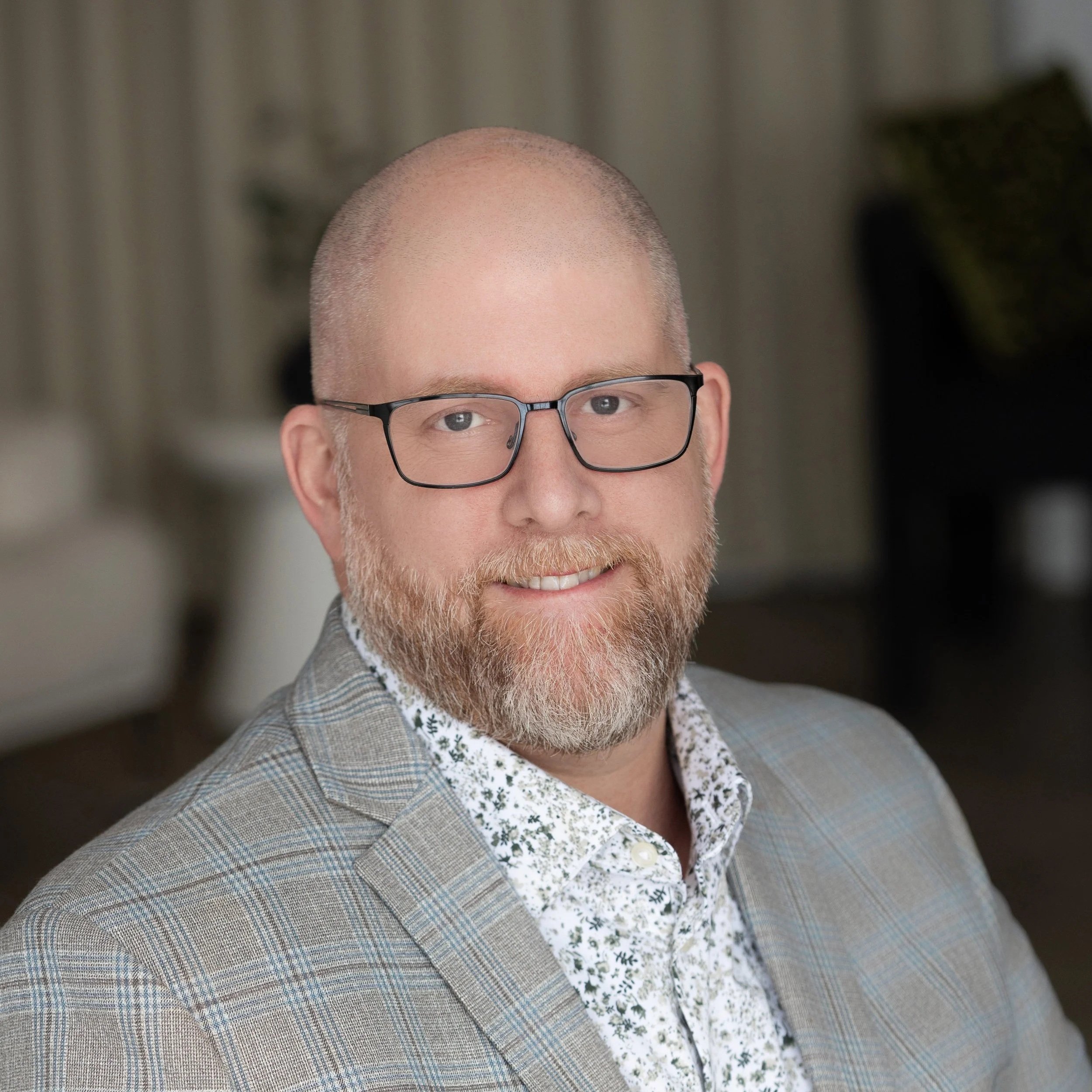 Close-up of a man with a bald head, glasses, and a beard wearing a blazer over a floral shirt, smiling in front of a blurred background.