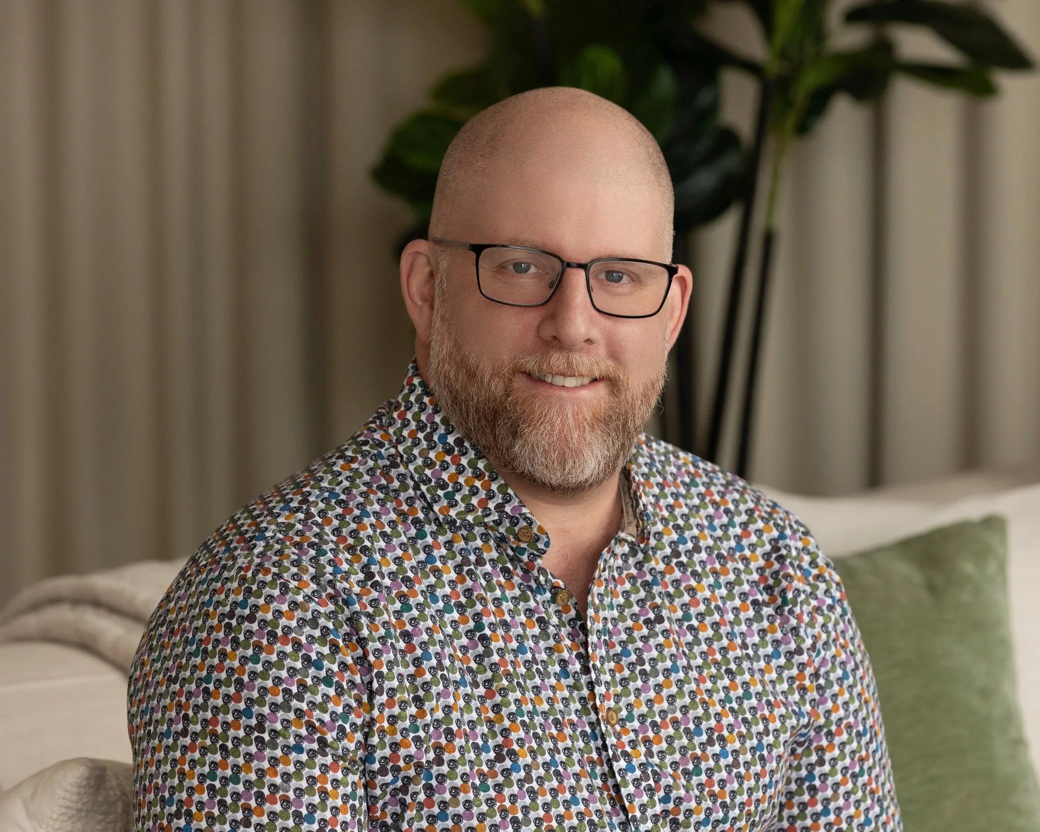 Scott Leibowitz with glasses, a beard, and a shaved head wearing a colorful, patterned shirt, sitting in a cozy indoor setting with a couch, a large plant, and curtains in the background.