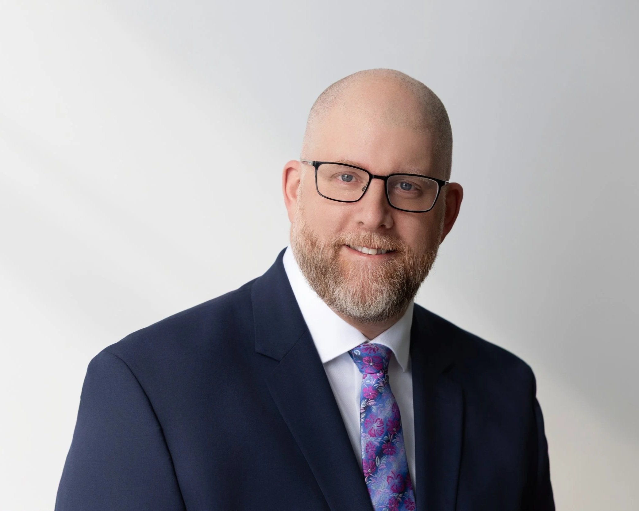 A man with a beard and glasses wearing a dark suit, white shirt, and a colorful floral tie, smiling in front of a plain white background.