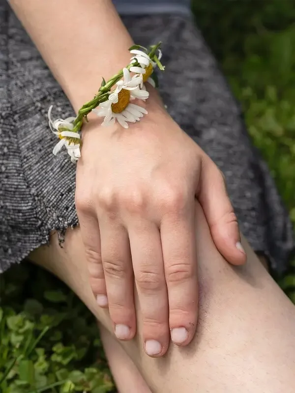 A person's hand resting on their knee, wearing a flower bracelet with daisies and other small white flowers, outdoors on grass.