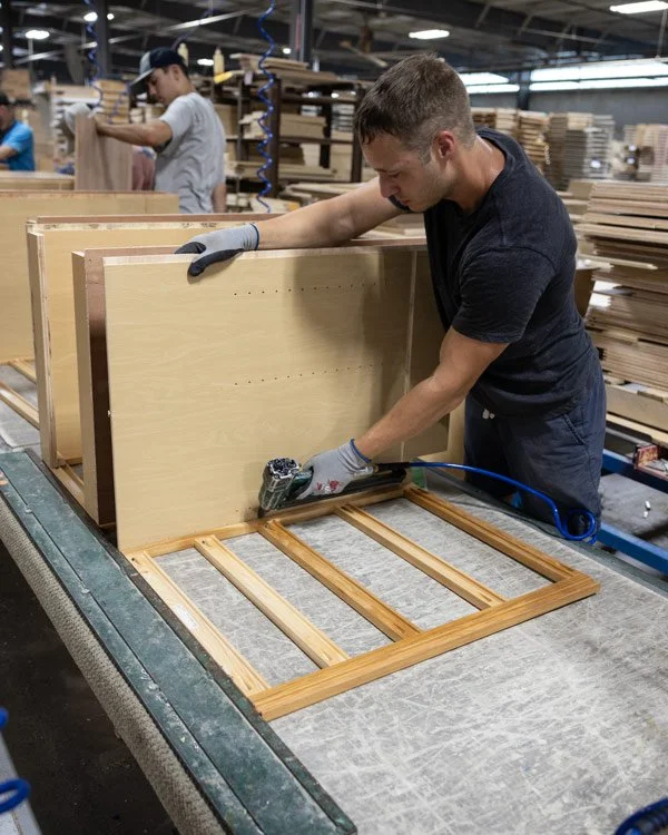 A worker using a power tool to assemble a wooden piece in a furniture manufacturing factory, with another worker in the background also working on wood.